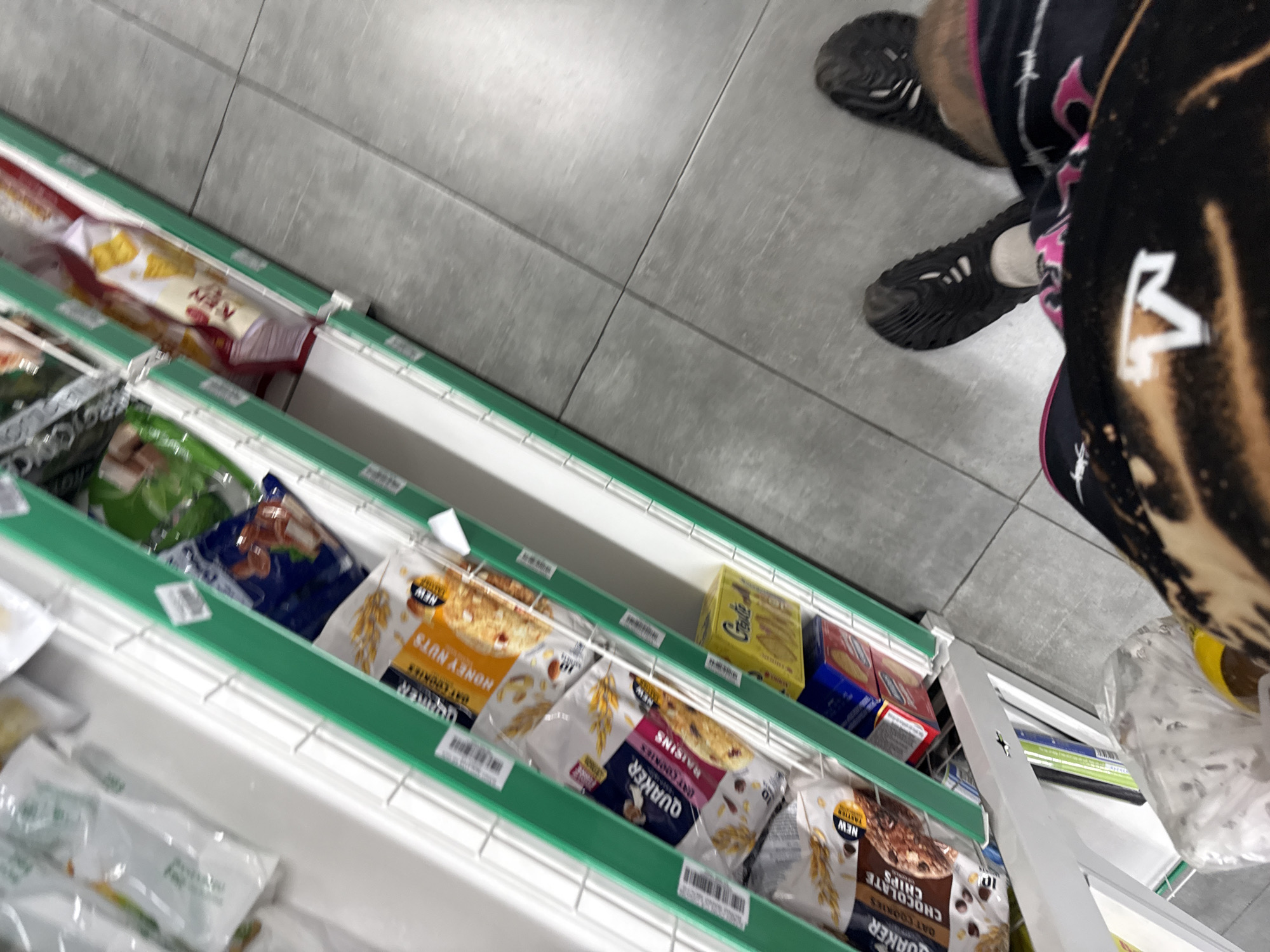 A shopper stands in the aisle of a supermarket. The aisle is filled with a refrigerated section of shelves containing food items, including cookies and snacks. Several packages of Quaker oat cookies are visible, along with boxes of Goute cookies. The shopper is wearing black shoes and clothing with pink and white detailing. A white plastic bag is in the lower right corner. The location is unknown.