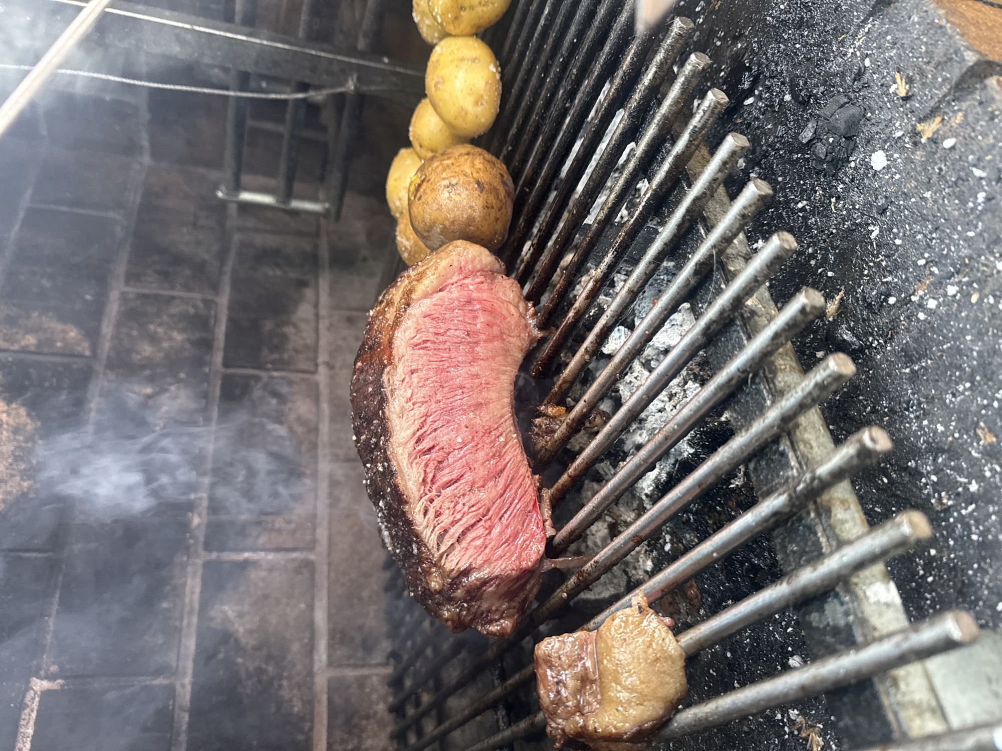 The image depicts a close-up view of a barbecue grill. On the grill, there is a medium-rare steak with a browned exterior. Also on the grill, a string of potatoes is being cooked. The grill is positioned over a brick and mortar structure, with smoke visible near the steak. The grill is positioned outdoors. Although not possible to determine solely from the image, this is potentially a barbecue in Barcelona, Spain.