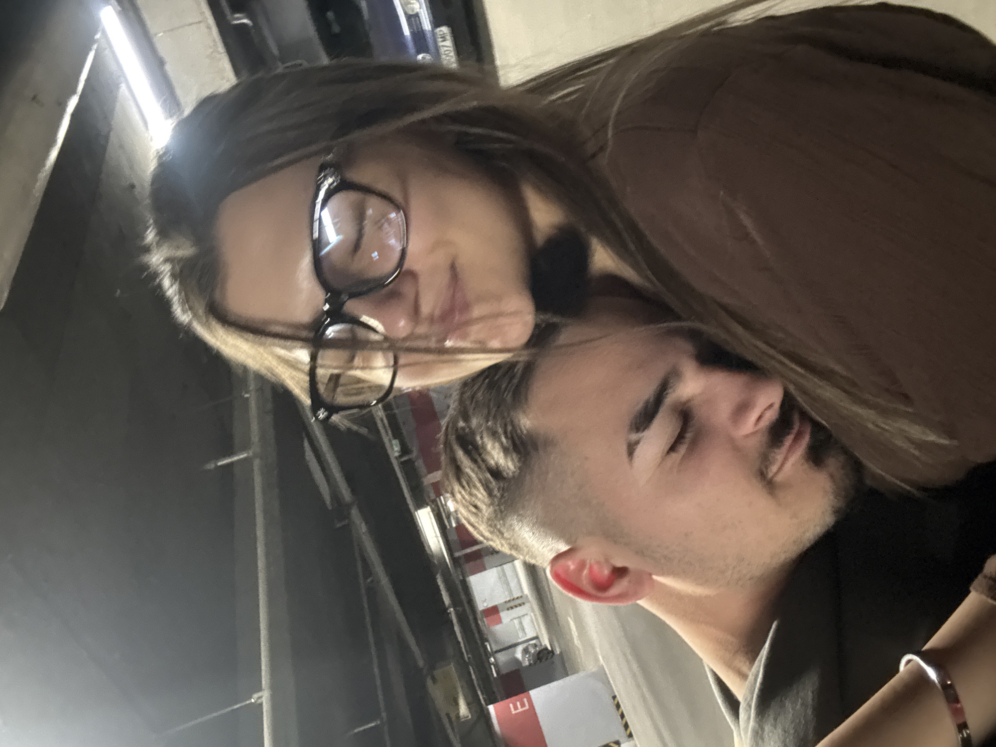A woman with glasses and a brown shirt is leaning her head on the head of a man. The man has short brown hair with stubble. The photo was taken in an indoor parking garage with fluorescent lighting. A silver bracelet is visible on the woman's wrist.