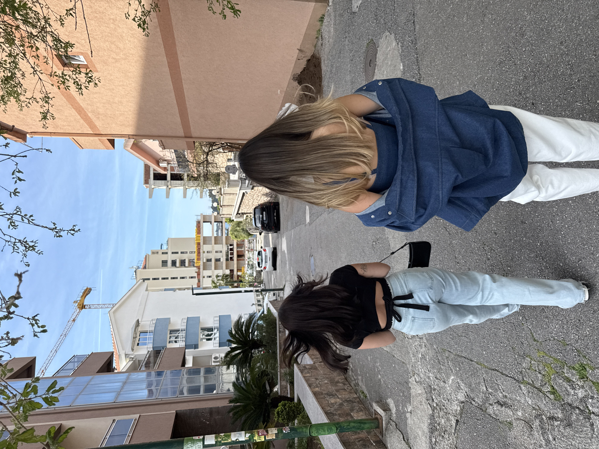 Two women stand on a city street, their backs to the viewer. The woman on the left has light brown hair, wears a denim jacket over a matching top, and white pants. The woman on the right has long dark hair, a black top tied in the back, light blue jeans, and carries a black purse. The street is flanked by buildings and palm trees. A crane is visible near one of the buildings. Several cars are parked on the street ahead. The sky is blue with few clouds.