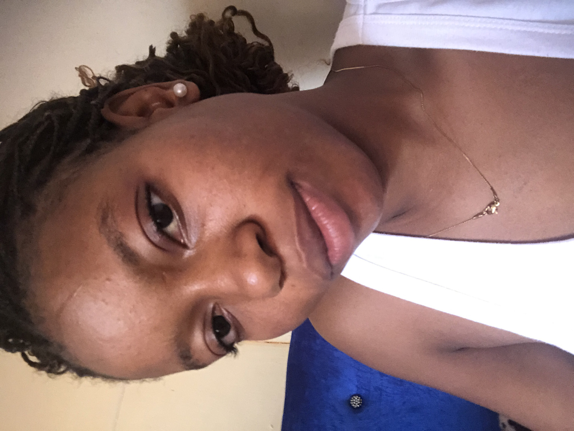 A person with dark skin, styled hair, and a pearl earring is visible in this close-up shot. They are wearing a white top and a delicate gold necklace. The background is ambiguous, with a mix of light beige and blue colors. The lighting appears to be bright and even. The location is unspecified.