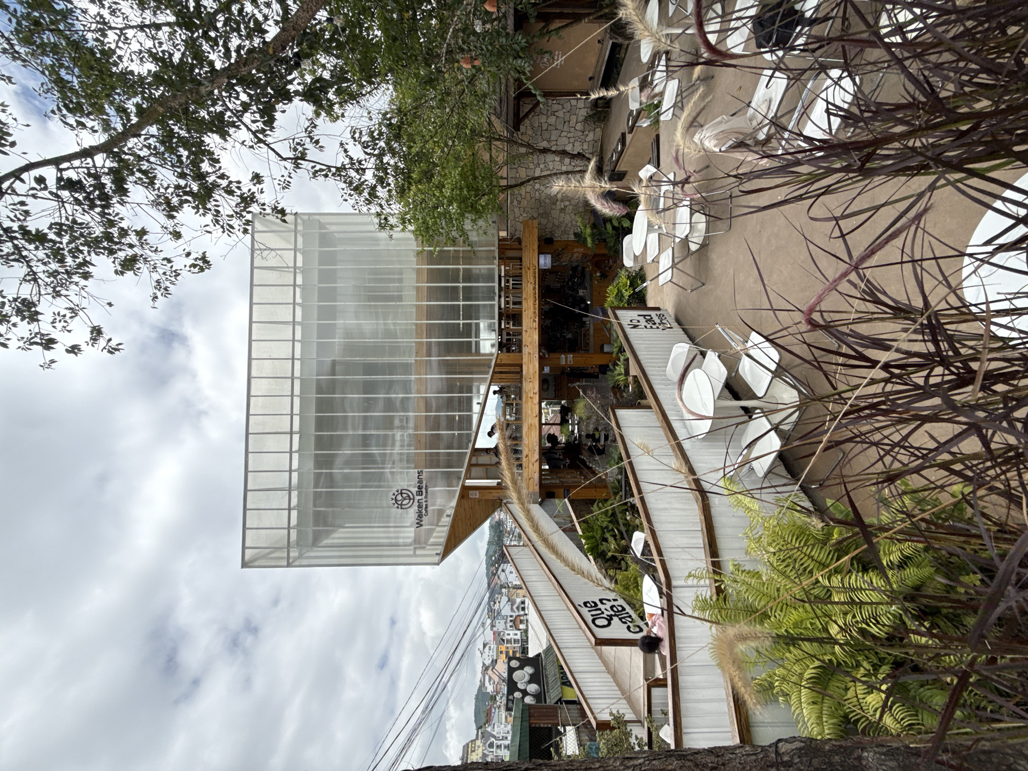 The image captures the exterior of Waken Beans Coffee & Roastery, a multi-level establishment situated in the city of Da Lat, Vietnam. The building features a modern, minimalist design with a transparent, ribbed facade on its upper level, contrasted with wooden accents and a stone base on the lower levels. An exterior staircase with white railings and signage that reads "Qué café?" and "No Fad Vibes" leads up to the building.
The scene is set outdoors with various seating areas, including white circular tables and chairs, scattered across a paved ground. Lush green ferns and tall, dark grass-like plants are visible in the foreground, adding a natural element to the urban landscape. In the background, a dense residential area with multi-colored houses climbs a hillside under a cloudy sky. The overall ambiance suggests a contemporary cafe with an inviting outdoor space.