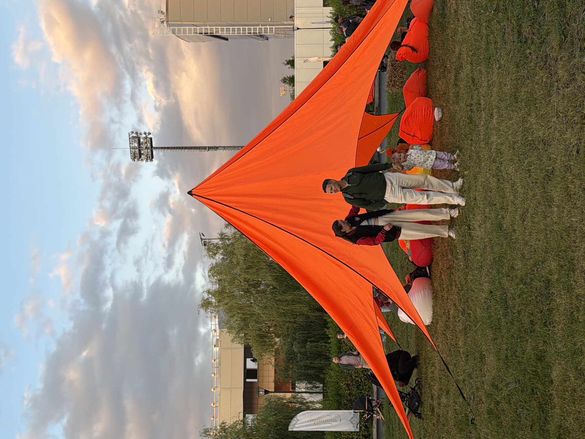 In an outdoor setting, an orange tent with black trim is pitched on a grassy field. A family, including a man, woman, and young child, stands near the tent. The man wears a green shirt and white pants. The woman wears a black jacket with white trim and light-colored pants. The child wears a patterned shirt and purple pants. Several large red beanbag chairs are scattered around the tent. In the background, there are trees, a building with a ladder, and a tall stadium light. The sky is partly cloudy with hints of pink and orange from the sunset.