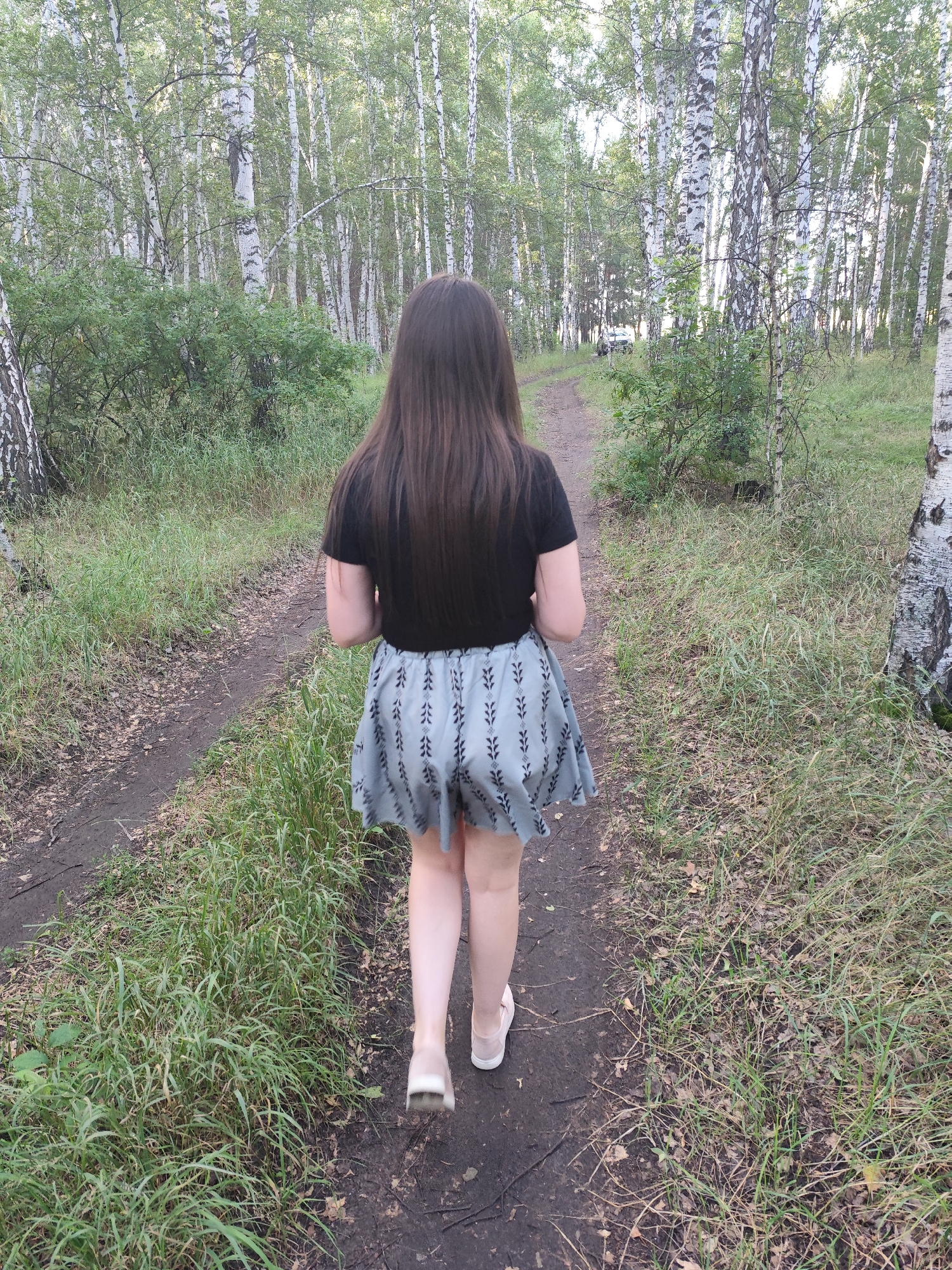 A young woman with long, dark hair walks away from the camera on a dirt path in a birch forest. She is wearing a black t-shirt and a light blue skirt with a pattern of dark leaves or branches. Her legs are bare, and she is wearing light-colored slip-on shoes. The path is flanked by tall birch trees with white bark and green leaves, and lush green grass and foliage. The sunlight filters through the trees, casting dappled shadows on the path and the surrounding vegetation. In the distance, on the path, a silver car is visible. The scene appears to be set during the daytime, possibly late afternoon or early evening, given the quality of light. The overall mood is serene and peaceful. There is no visible text in the image.