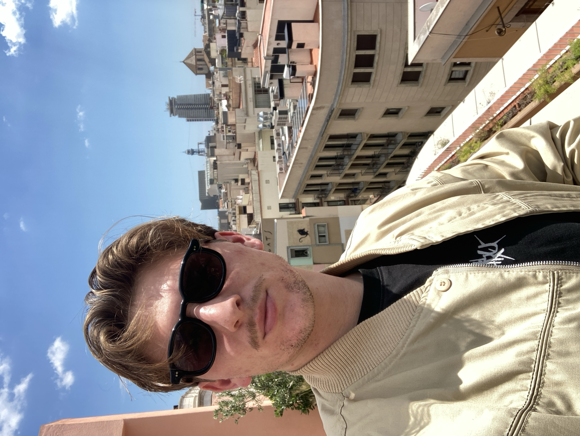A slightly rotated medium eye-level shot depicts a man with light brown hair wearing dark sunglasses, a tan jacket, and a black shirt, positioned against a cityscape background under a bright blue sky with scattered clouds. The man has fair skin and stubble. The cityscape includes a variety of buildings in neutral tones, with a tall, modern tower in the distance. A small green tree is visible in the lower left of the frame. This image likely takes place in Barcelona, Spain, as the buildings and tower are characteristic of its architecture.