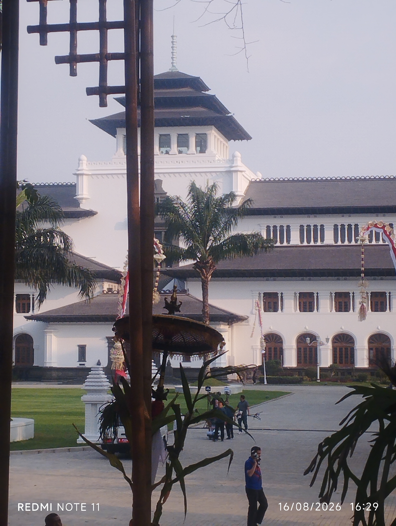 This image depicts Gedung Sate, a prominent historical landmark in Bandung City, Indonesia. The architectural style of the building is evident, with its white facade, dark tiled roofs, and ornate window designs. A tiered tower with a spire rises from the center of the building. The foreground is partially obstructed by dark vertical wooden poles and lush green foliage, suggesting a viewing perspective from within a garden or park area. In the middle ground, several individuals are visible, some engaged in what appear to be outdoor activities or possibly a filming setup with equipment. The grounds are well-maintained with green lawns and paved pathways. The sky is overcast and hazy, indicating a bright but diffused light. The overall impression is a serene, historical setting with signs of contemporary activity.