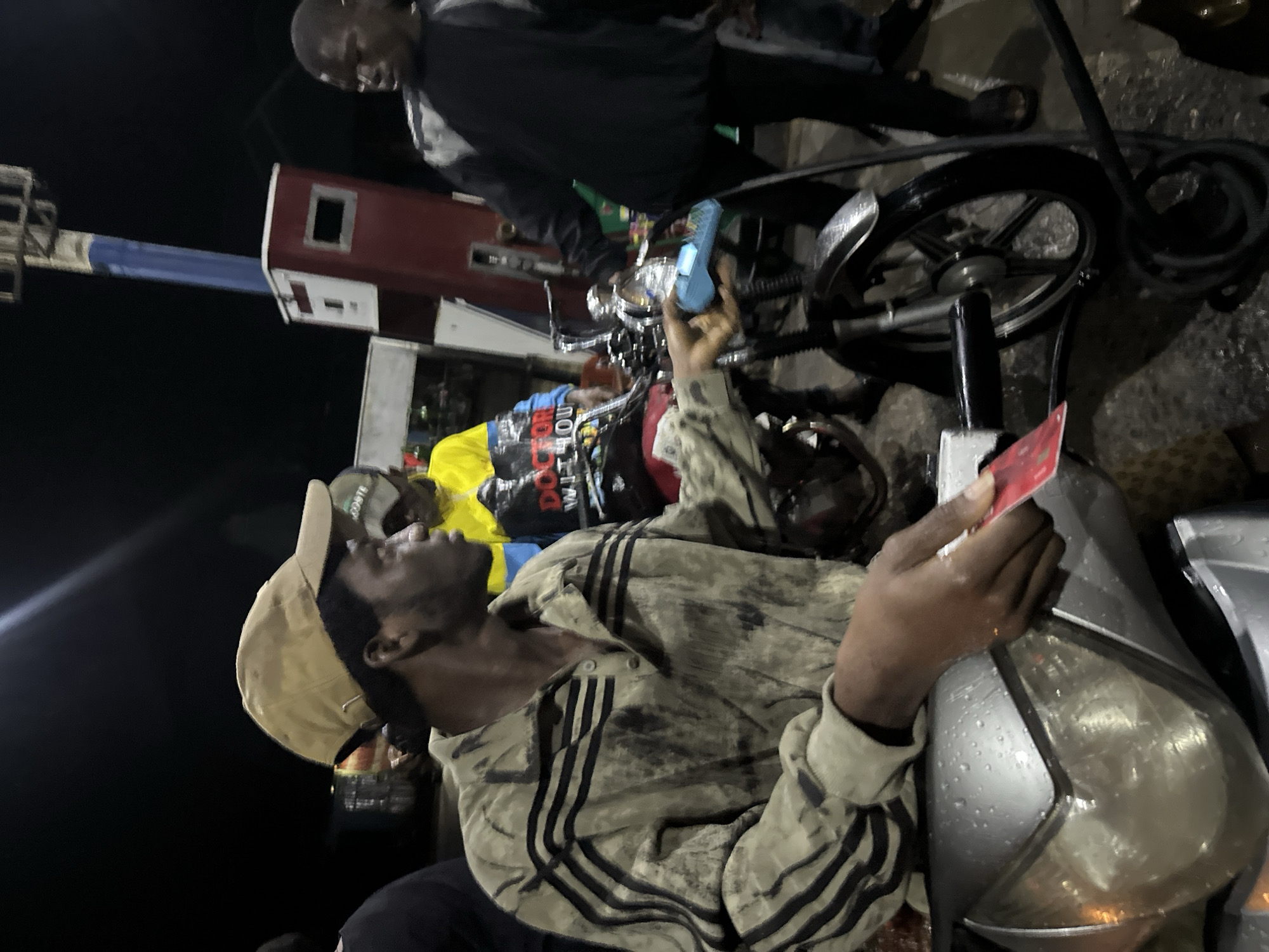 At a gas station in Ikot Ekpene, Nigeria, a man wearing a tan baseball cap, and a striped collared shirt is seated on a wet, gray motorcycle. He is holding a red card, potentially a credit card or similar payment method. Beside the motorcycle, another man, wearing a dark shirt, is standing near a bicycle, holding a small blue object. Behind them, there is a gas pump with a red and white structure. The scene appears to be at night, as indicated by the dark background.