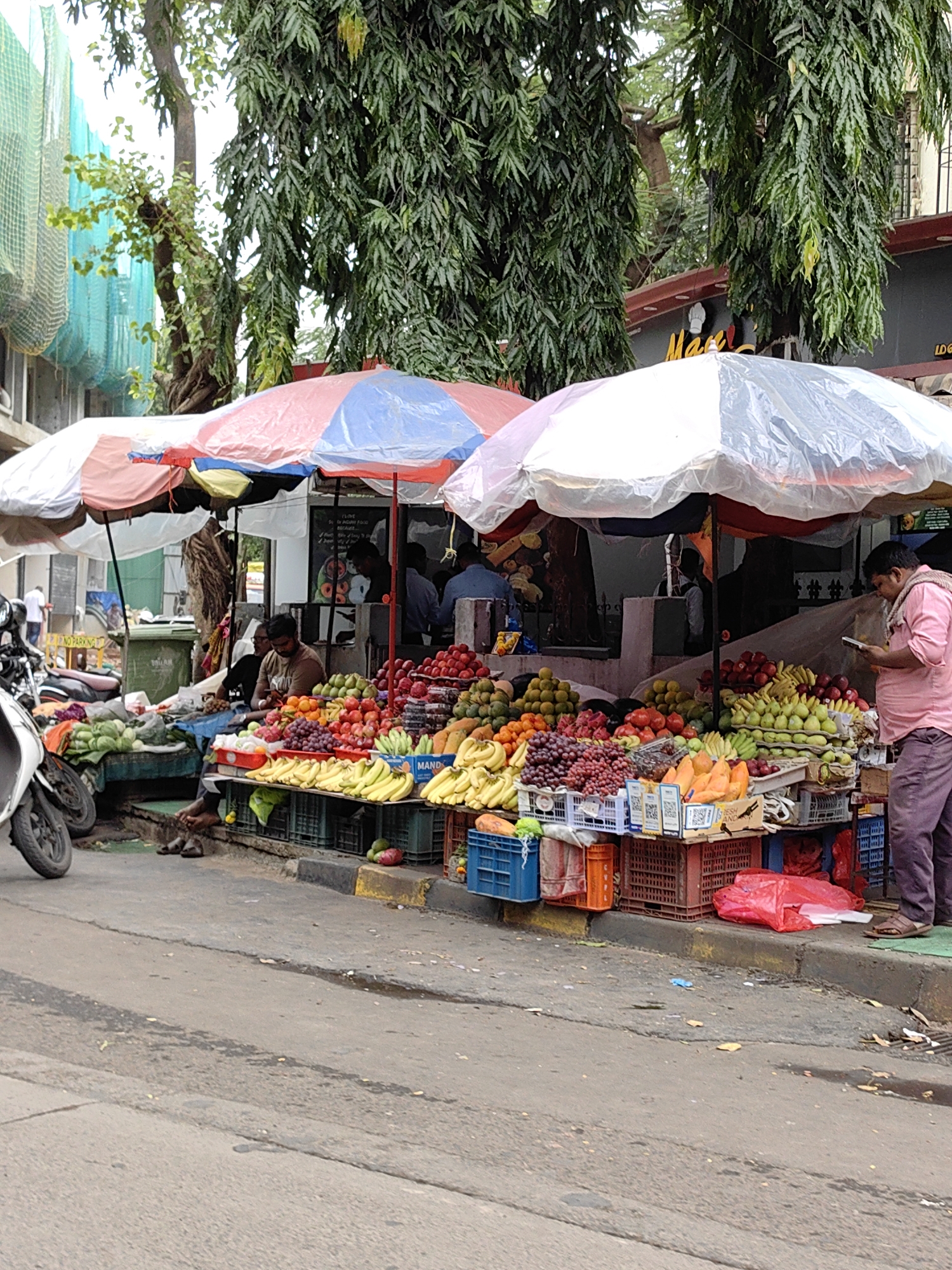 An outdoor fruit and vegetable stall in Mumbai, India, is depicted under large, colorful umbrellas. The stall displays an abundant variety of produce, including bananas, grapes, apples, oranges, and other fruits and vegetables, arranged in crates and on tables. Several individuals are present: a man in a pink shirt is using a mobile phone while standing near the produce, another man in a brown shirt is seated and appears to be attending to the stall, and other figures are partially visible in the background. The stall is set up along a roadside, with a street in the foreground and a building with trees behind it. A motorcycle is parked to the left of the frame. The scene suggests a typical day at a bustling local market in the city.
