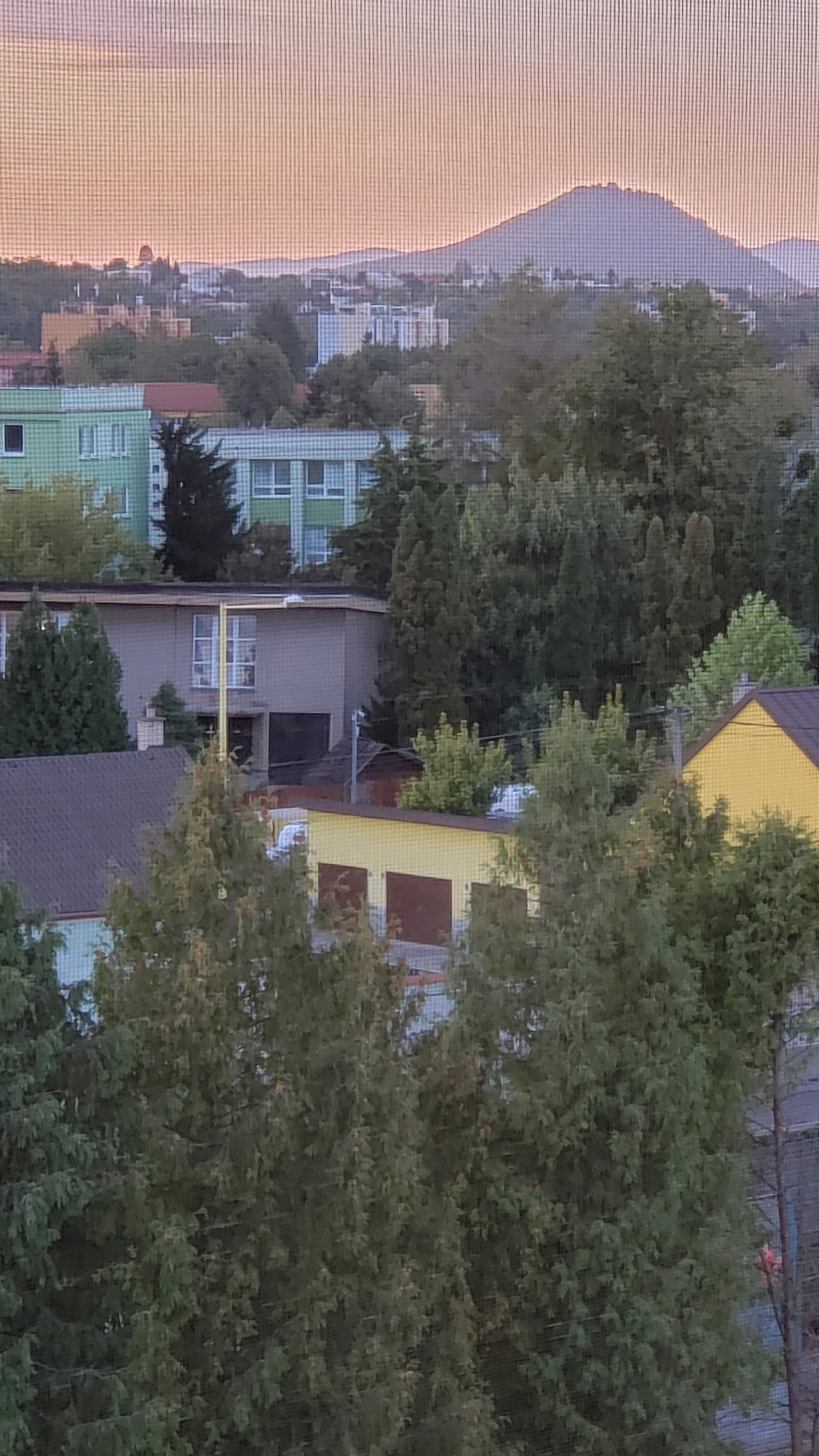 The image shows a view of the city of Prešov, Slovakia, taken through a window screen. In the background, a large mountain range is visible under a hazy sky. The middle ground is filled with buildings of various sizes and colors, including apartment blocks with green and white facades, and smaller structures with yellow and brown roofs. Lush green trees are interspersed throughout the urban landscape, some appearing to be mature evergreens. The foreground is dominated by the dense foliage of trees, which obscures some of the lower buildings. The overall impression is a suburban or urban setting with significant green spaces, likely captured during daylight hours given the illumination.