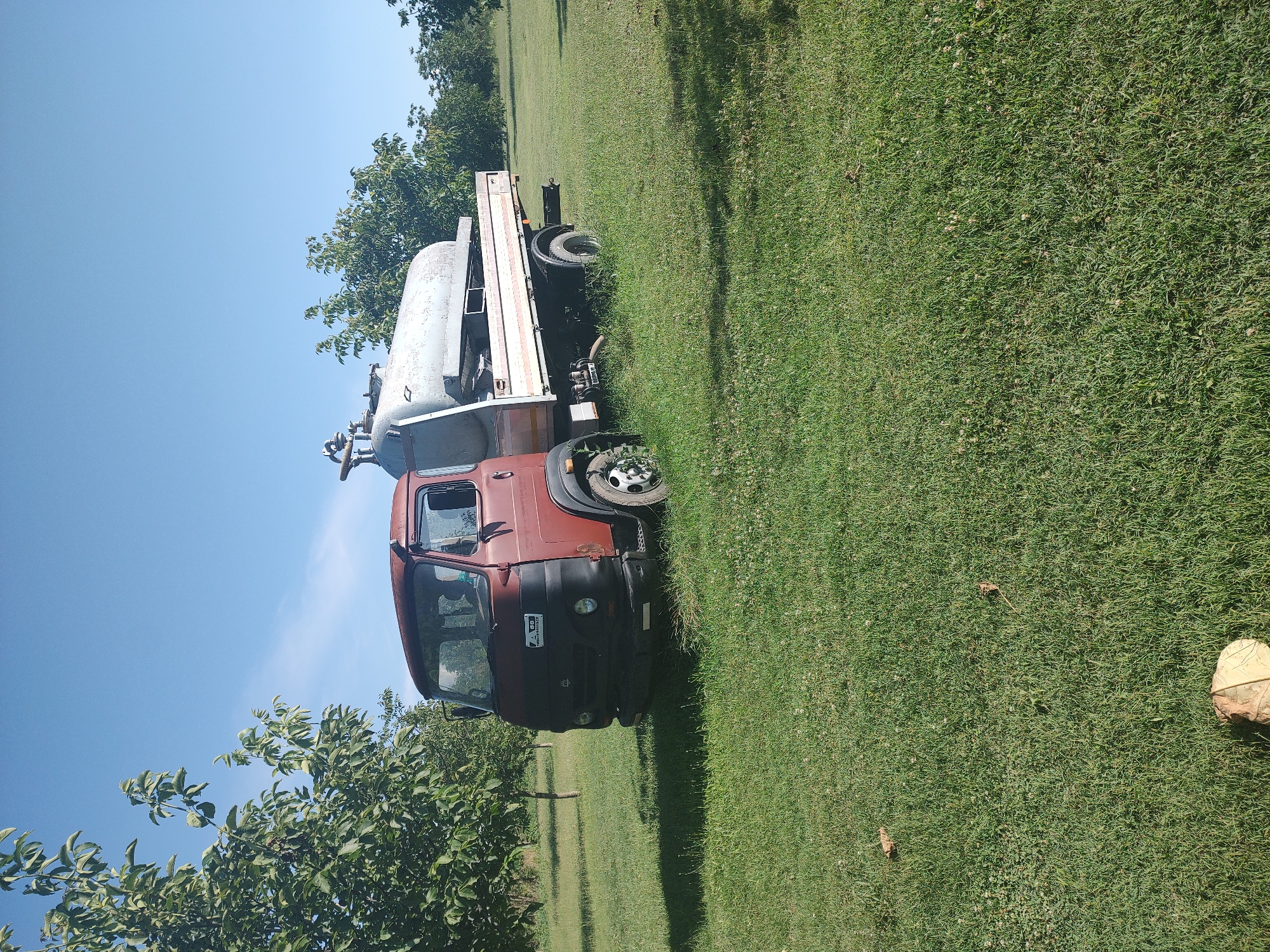 An aged, rust-colored truck with a large, silver tank mounted on its flatbed is parked on a grassy field under a clear blue sky. The truck appears to be in a rural or orchard setting, with trees visible in the background and to the left of the vehicle. The tank has some visible piping and valves. The truck's front grille is black, and there is a sticker on the driver's side door. The grass is green and somewhat uneven, with some fallen leaves scattered on the ground. The scene suggests a sunny day, possibly in the city of Kochmar, Bulgaria, given the context provided, implying a location within the agricultural landscape of the region.