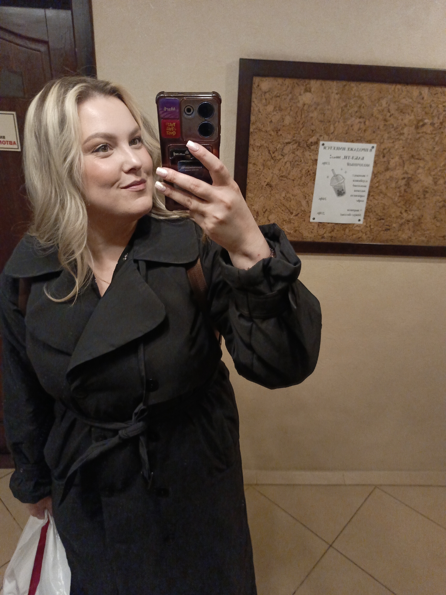 A young woman with blonde hair and light makeup is captured in a selfie taken indoors, likely in a hallway or common area in Chaikovsky, Russia. She is wearing a black trench coat, tied at the waist, and a delicate gold necklace. Her manicured nails, painted with glittery polish, are visible as she holds a smartphone with a distinctive dark case featuring several stickers or inserts, some of which show reversed text like "Mare" and "One."
The woman is looking slightly to her right, with a soft smile. Behind her to the right is a large cork bulletin board with a dark wooden frame, pinned with several notices. One prominent, reversed notice features a drawing of a drink cup and partially visible Russian text, including words that appear to be "РОЗЫГРЫШ" (RAFFLE) and "ВЫИГРЫШ" (WIN). To her left, a dark wooden door is visible, with some reversed text on a sign that reads "МОТВА" (MOTVA). The walls are a neutral beige color, and the floor is tiled with light-colored squares and darker grout lines. She holds a white shopping bag with a red stripe in her left hand. The overall lighting suggests an indoor setting, and the activity is clearly a mirror selfie.