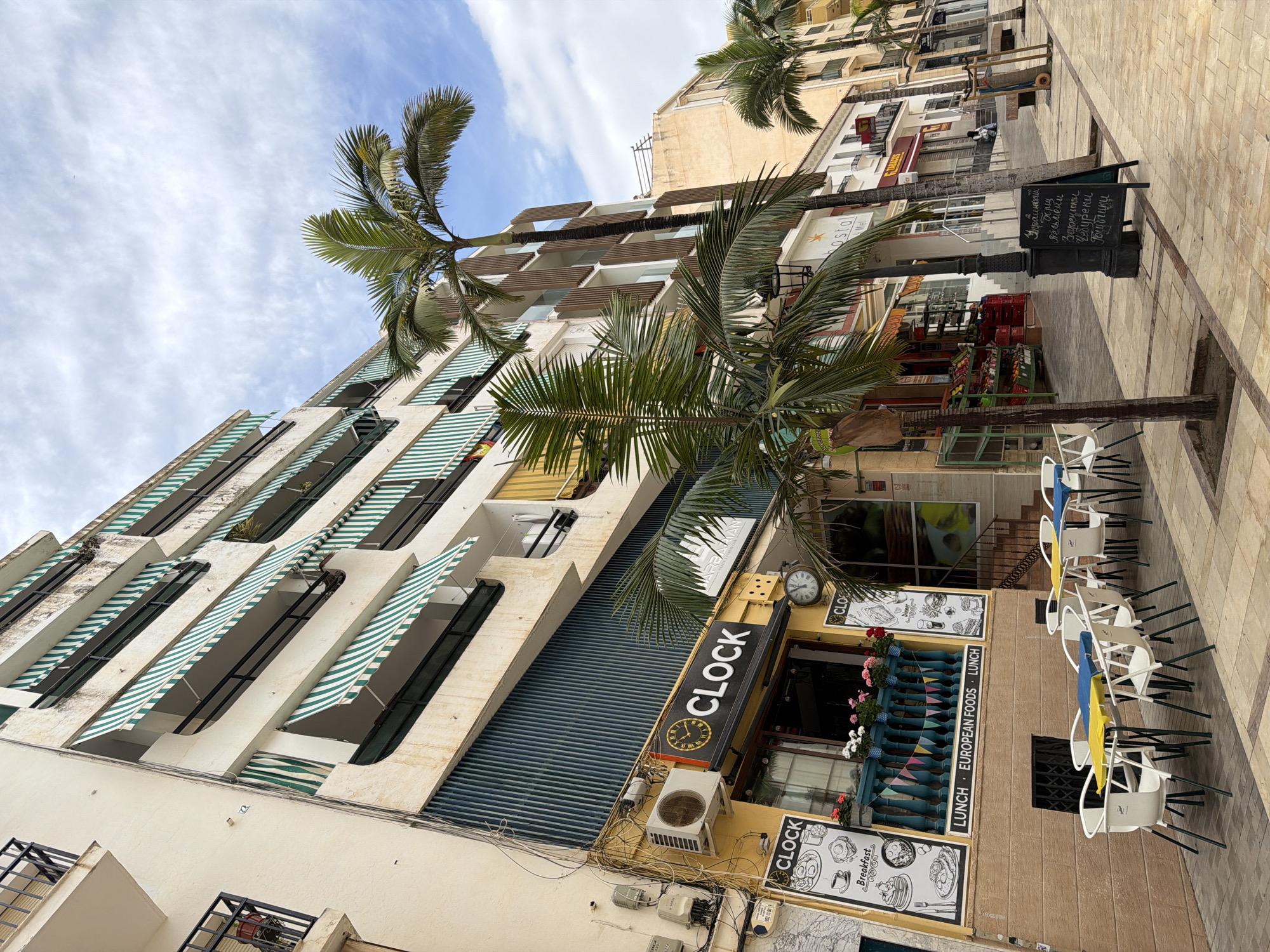 The image shows a street scene in Arroyo De La Miel-Benalmadena Costa, Spain. In the foreground is a cafe named "CLOCK," with an awning, menu board, and outdoor seating consisting of white chairs with colored (yellow and blue) fabrics. The cafe's exterior is decorated with flowers, and a clock is visible above the entrance. Adjacent to the cafe are tall buildings, one with green and white striped awnings. Palm trees are positioned along the street and partially obscure the view of the buildings. A black board with Russian menus is partially visible and near the cafe. The environment is typical for a Mediterranean coastal city, with a mix of commercial and residential buildings and the presence of palm trees. The sky is partially cloudy.