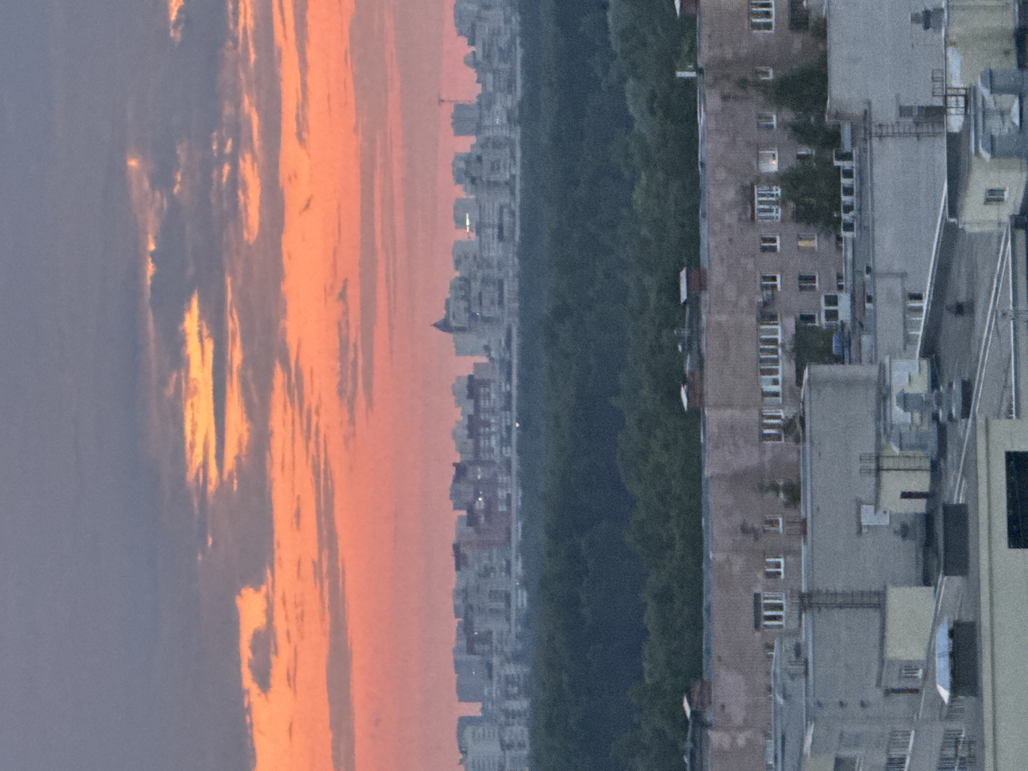 The image displays a cityscape during either sunrise or sunset, evidenced by the vibrant colors in the sky. The photo appears to have been taken in landscape orientation and then rotated 90 degrees clockwise.
In the left portion of the image (when oriented correctly), the sky transitions from a soft blue-grey on the far left to striking oranges, pinks, and yellows on the right, where the sun's light is catching scattered clouds.
The right portion of the image reveals an urban landscape. A dense band of dark green trees runs across the middle ground, likely indicating a park or wooded area. Beyond this green space, a distant city skyline is visible, comprised of numerous grey multi-story buildings that appear as silhouettes against the colorful sky. A few lights are visible in these distant structures.
In the foreground, closer to the viewer, are more detailed buildings, characterized by multiple windows and balconies. These structures are predominantly grey and brown, suggesting an older architectural style. No people are visible, and no specific activities are taking place. The scene is static, capturing a moment of natural beauty over an urban environment. There is no visible text. The time of day is clearly either early morning or late evening, and the weather appears calm with some cloud cover.