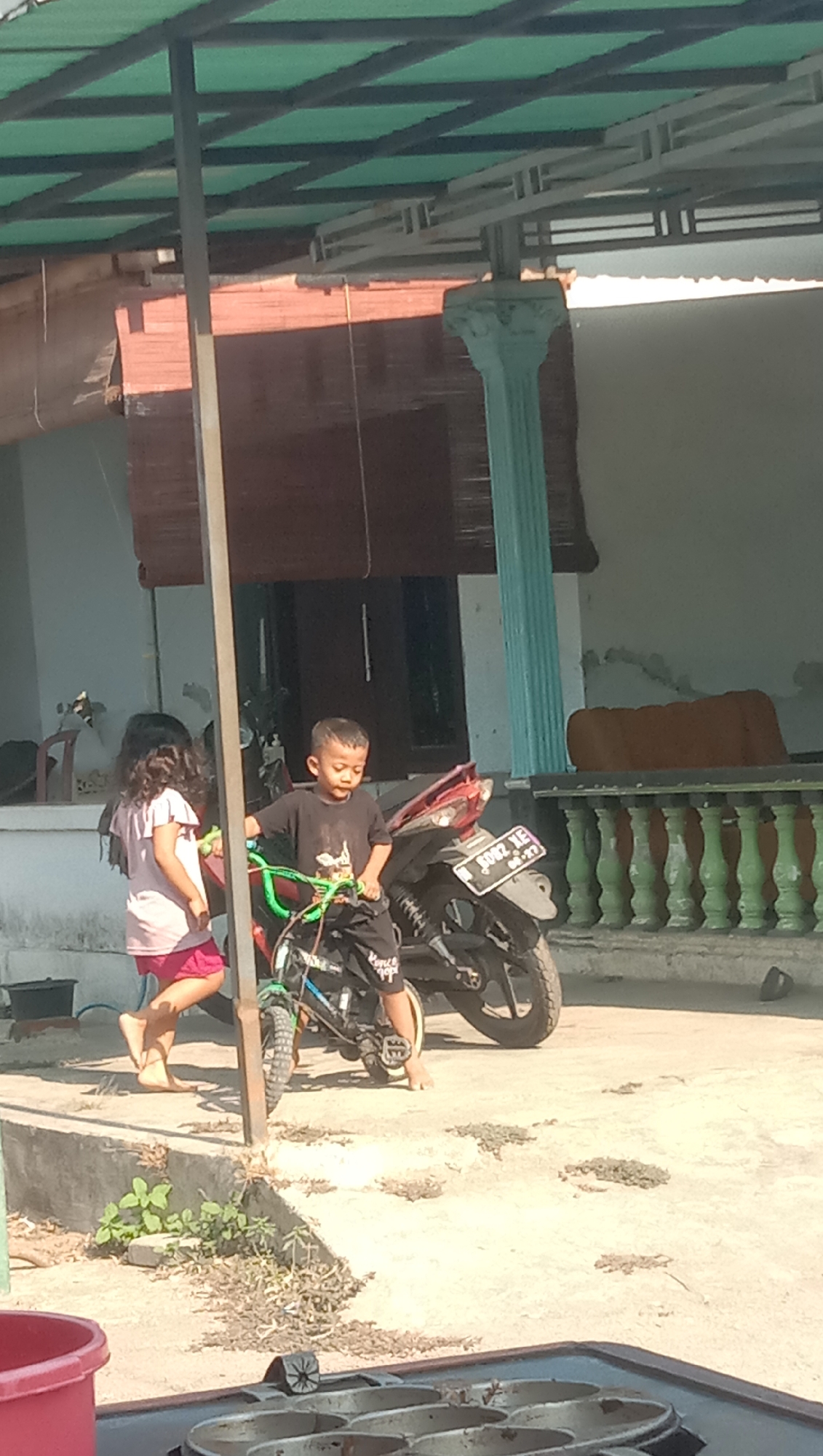 In Sidogiri, Indonesia, two children are outdoors near a building with a green roof. A young boy in a black t-shirt and black pants stands with his hands on the handlebars of a green bicycle. Next to him, a girl in a pink t-shirt and pink shorts walks away from the bicycle. A motorcycle with a license plate is parked behind the children. The ground is paved with concrete and has patches of dry grass. The background shows a building with a blue pillar and a brown woven screen. The scene is illuminated by bright sunlight.