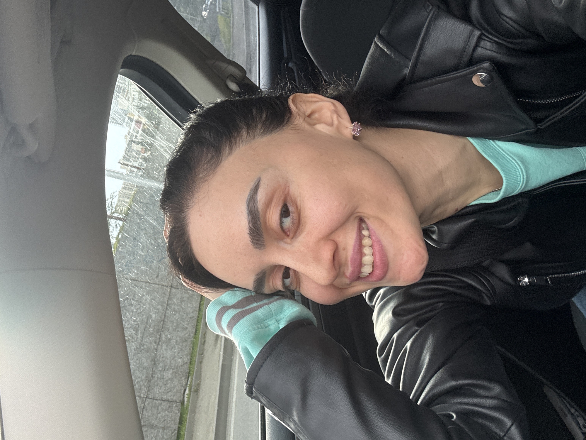 A young woman with dark hair and prominent eyebrows is captured in a headshot inside a car, looking directly at the camera with a broad, toothy smile. Her head is tilted to her right, and her left arm is bent, with her hand near her head. She wears a black leather jacket over a light blue long-sleeved top with horizontally striped cuffs in light blue and grey. A small, pink stud earring is visible in her right ear, and a thin silver chain adorns her neck.
The setting is the interior of a vehicle, with parts of the car's window frame, an A-pillar, and a dark seatbelt visible. Bright natural light illuminates the scene, indicating daytime. Through the car window, which has visible streaks, a blurry urban or suburban background can be discerned, including suggestions of buildings or structures and some greenery. The woman's expression conveys a happy and cheerful mood. There is no visible text in the image. The context for this photograph is Tbilisi, Georgia.