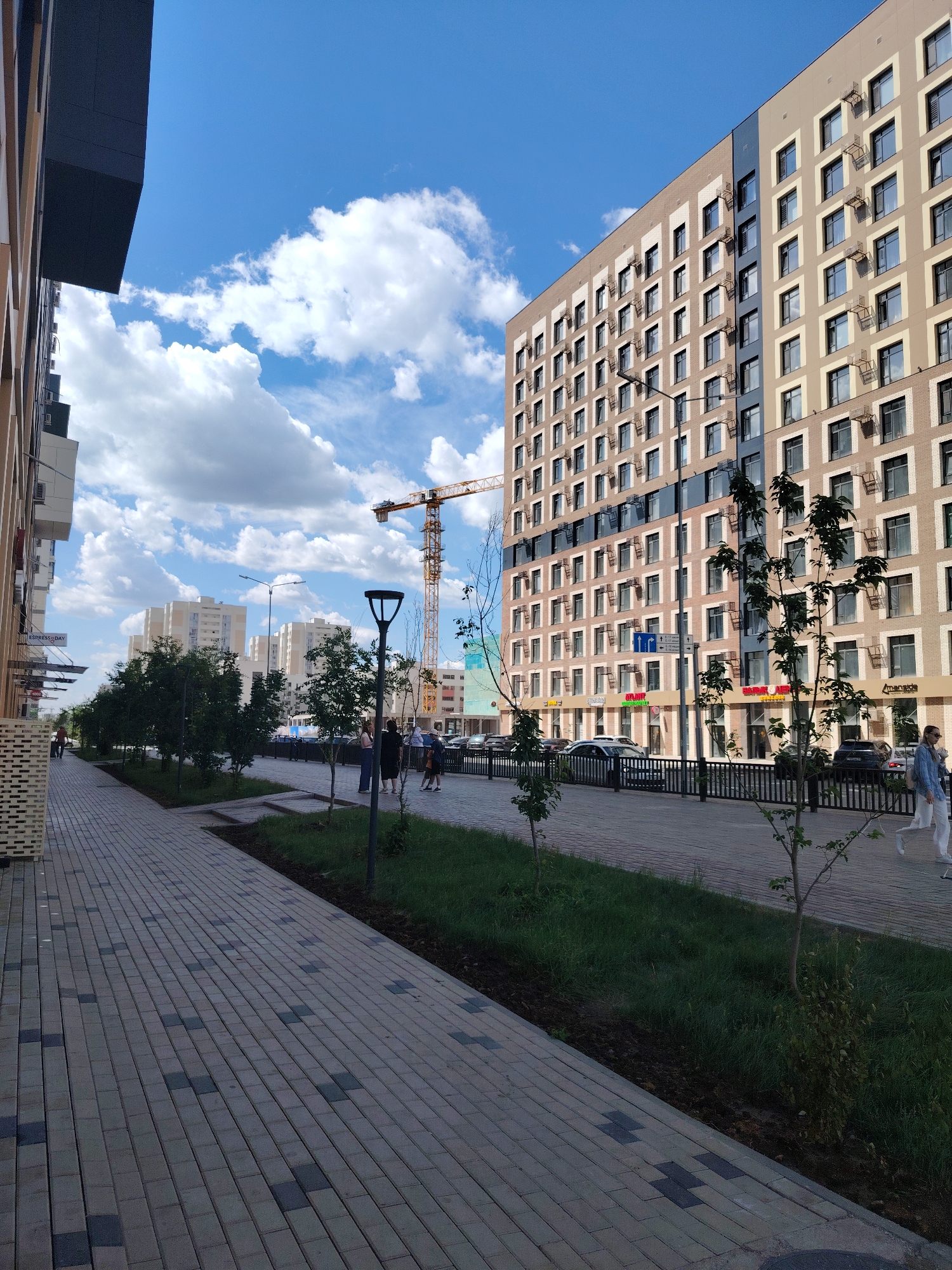 This media file depicts a modern urban scene in Astana, Kazakhstan, under a bright, partly cloudy sky. The image captures a wide pedestrian walkway paved with interlocking bricks in a grey and dark grey pattern, leading the viewer's eye towards a row of contemporary apartment buildings. On the left, a building with dark grey and beige facade elements is partially visible, extending upwards. On the right, a tall, multi-story apartment building with a beige and light brown facade dominates the view. This building features numerous windows, each framed by a beige border, and air conditioning units mounted outside many of them. A section of the building is accented with dark blue panels.
Along the right side of the walkway, a black metal fence separates the pedestrian area from a road where several cars are parked and driving. Beyond the fence, a strip of lush green grass and young trees line the street, with more mature trees planted along the pedestrian path. A tall street lamp with a black finish stands on the walkway, and further in the background, a large yellow construction crane is visible, suggesting ongoing development in the area.
Several people are casually walking along the pathway, some in small groups, indicating a normal day. The lighting suggests it is daytime, likely mid-morning or afternoon, with the sun casting shadows. The overall atmosphere is one of urban development and everyday life.
Visible text includes a sign that appears to read "ESPRESSO DAY" on the building to the left, and other signs on the ground floor of the apartment building on the right, some of which are difficult to decipher but appear to be shop names. The weather appears pleasant, with fluffy white clouds scattered across a clear blue sky.