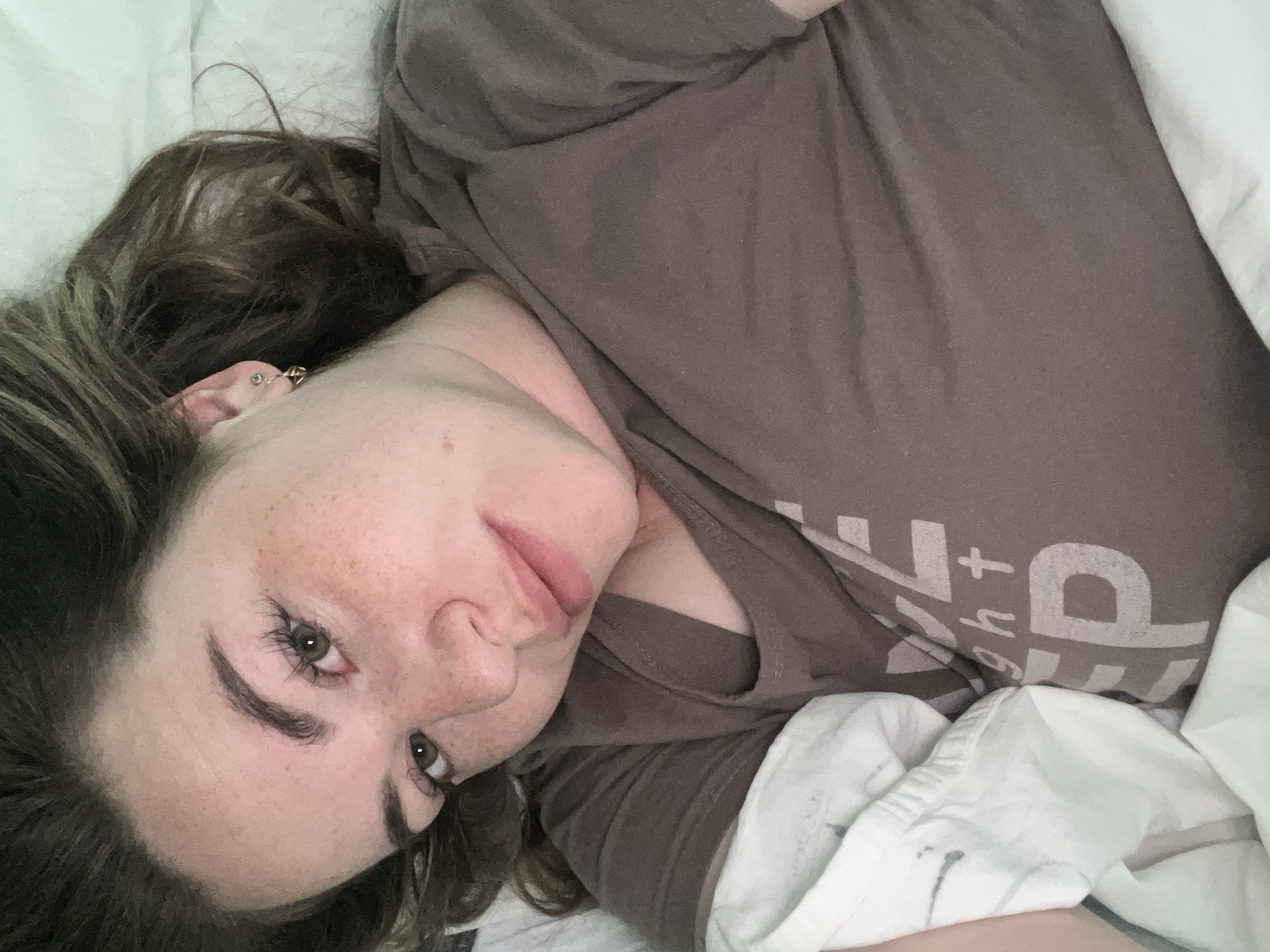 A woman lies in a bed with white bedding. She wears a brown t-shirt. She has light skin, brown hair, and freckles. The image provides no information to suggest the location is in Chaikovsky, Russia, or anywhere else specifically.