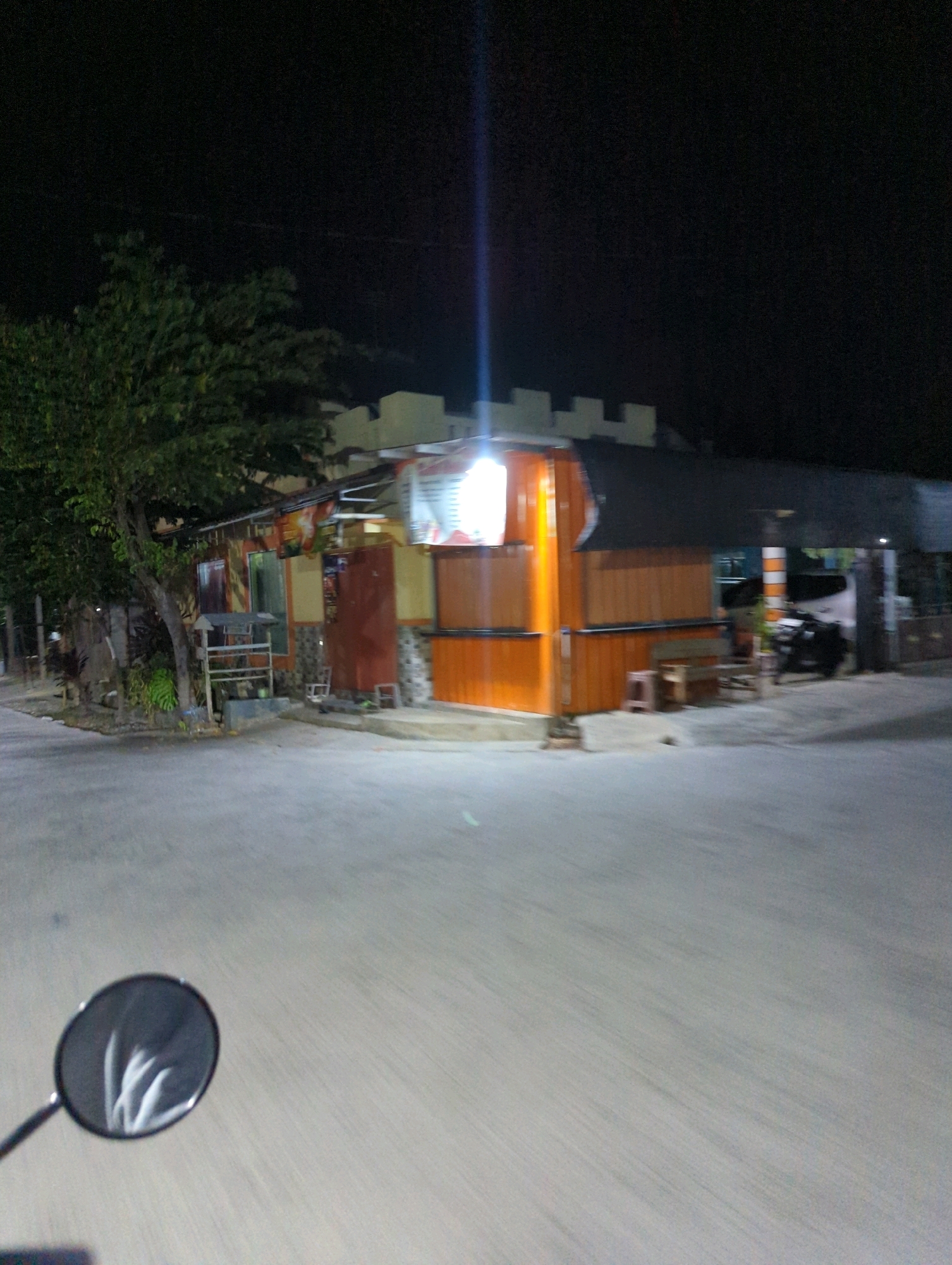The image depicts a street scene at night in Senggrong, Indonesia. A small building, likely a shop or food stall, is illuminated by artificial lights. The building has an orange facade with wooden paneling and a dark roof. Several windows and doors are visible. In front of the building, there are some chairs and a bench, suggesting a seating area for customers. A tree with dense foliage is situated to the left of the building, casting shadows. A motorcycle is parked in front of the building to the right, and a white vehicle is partially visible behind it. The street in the foreground appears to be paved and empty, with motion blur indicating movement. A motorcycle's rearview mirror is visible in the lower left corner of the image, reflecting the ambient light and possibly the surrounding environment. The sky is dark, characteristic of nighttime.