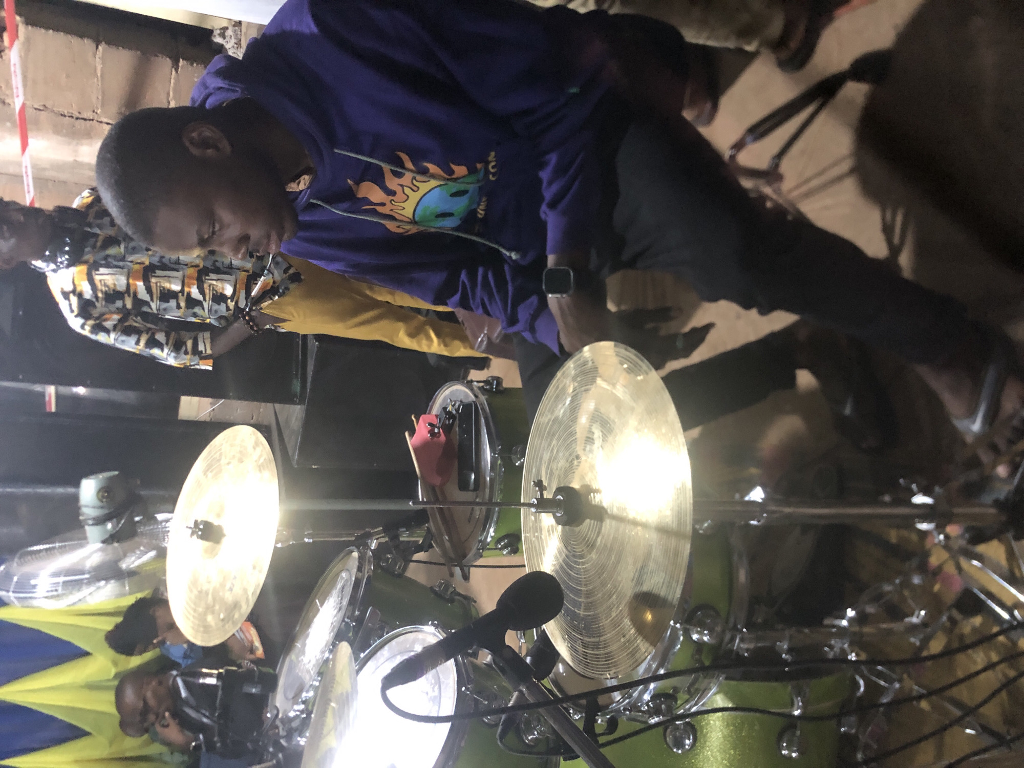 In Obosi, Nigeria, a man in a purple hoodie is playing a green drum set. Several other people are in the background.