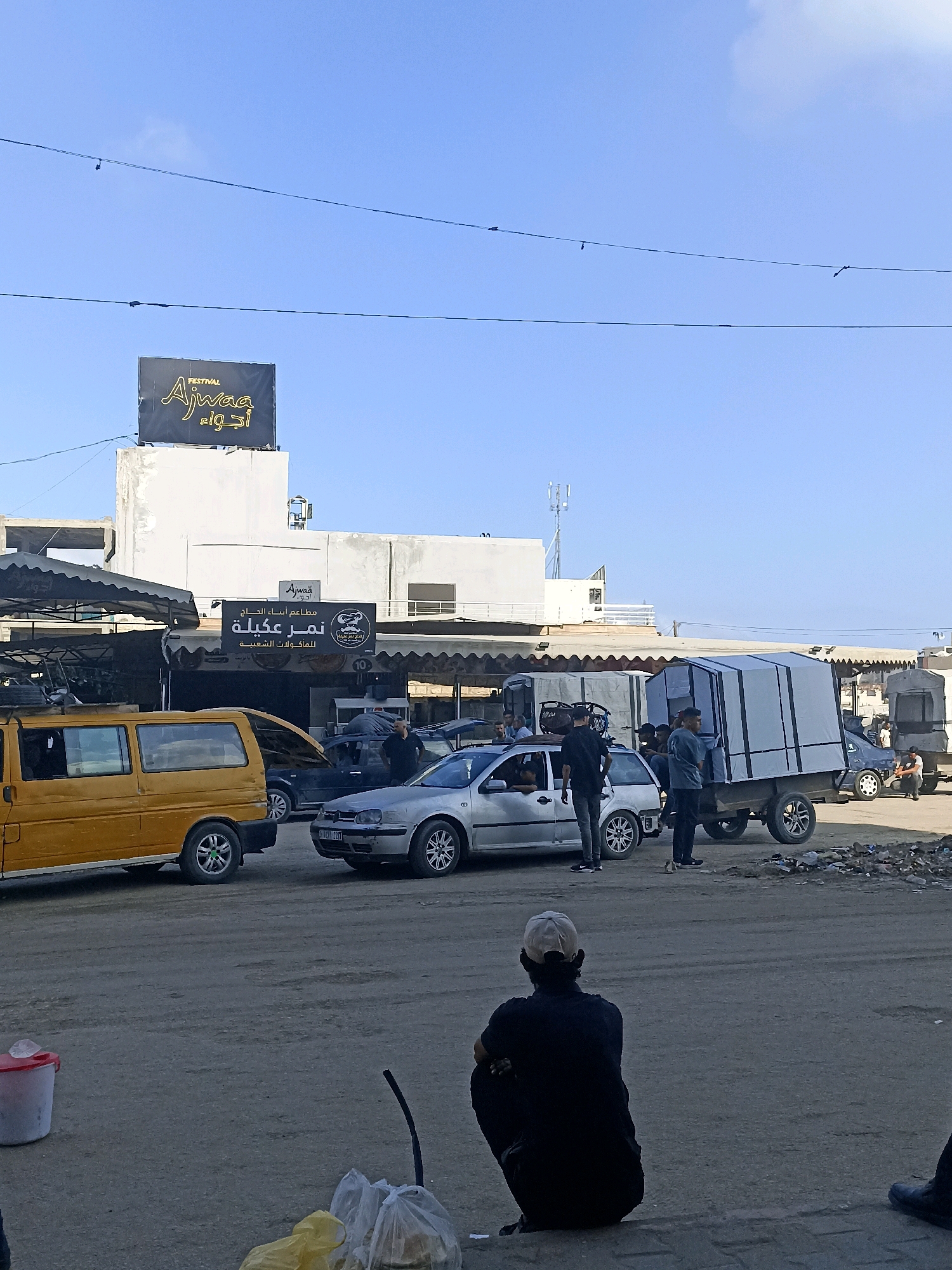 The image depicts a street scene in Gaza City, Palestinian Territories. In the foreground, a person wearing a dark shirt and a light-colored baseball cap is crouched down, facing away from the camera. Several vehicles are visible, including a yellow van on the left, a silver Volkswagen Golf, and a flatbed trailer with a white canopy. A group of men are standing around some of the vehicles, appearing to be engaged in activity related to loading or unloading the trailer. In the background, buildings are visible, one of which has a sign with "FESTIVAL Ajwaa" written on it in English and Arabic. Another sign below it reads "مطعم ابناء الحاج" (Haj's Sons Restaurant). The ground is paved, and there is some debris scattered in the area. The sky is clear and blue.