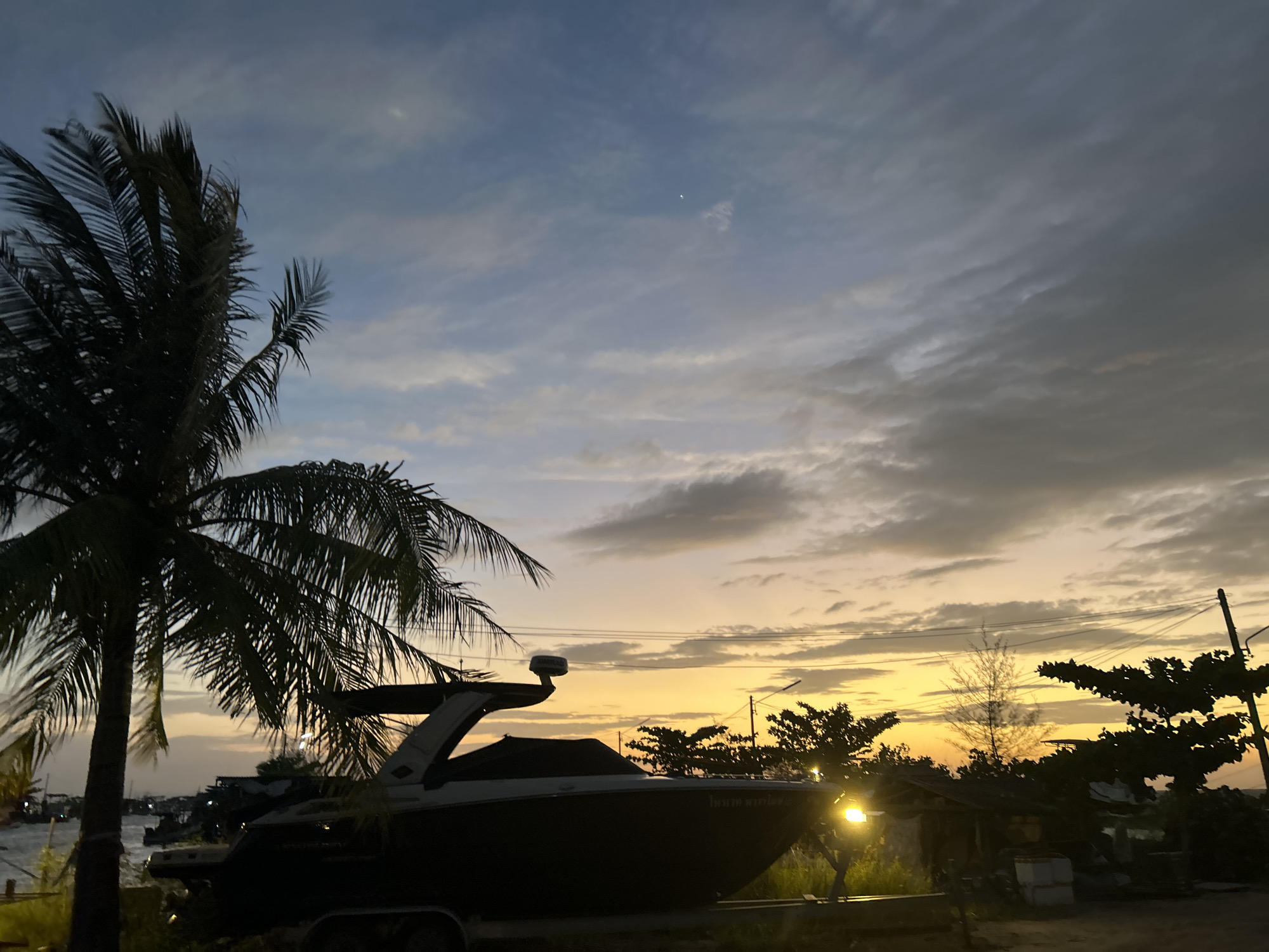 A boat is parked on a trailer in front of a palm tree during sunset on Ko Pha-ngan District, Thailand. The sky is filled with clouds and the sun is setting, casting a warm glow. There is a body of water in the background with some buildings and boats. The boat is dark in color and has a cover over part of it. There is a light on in the background.