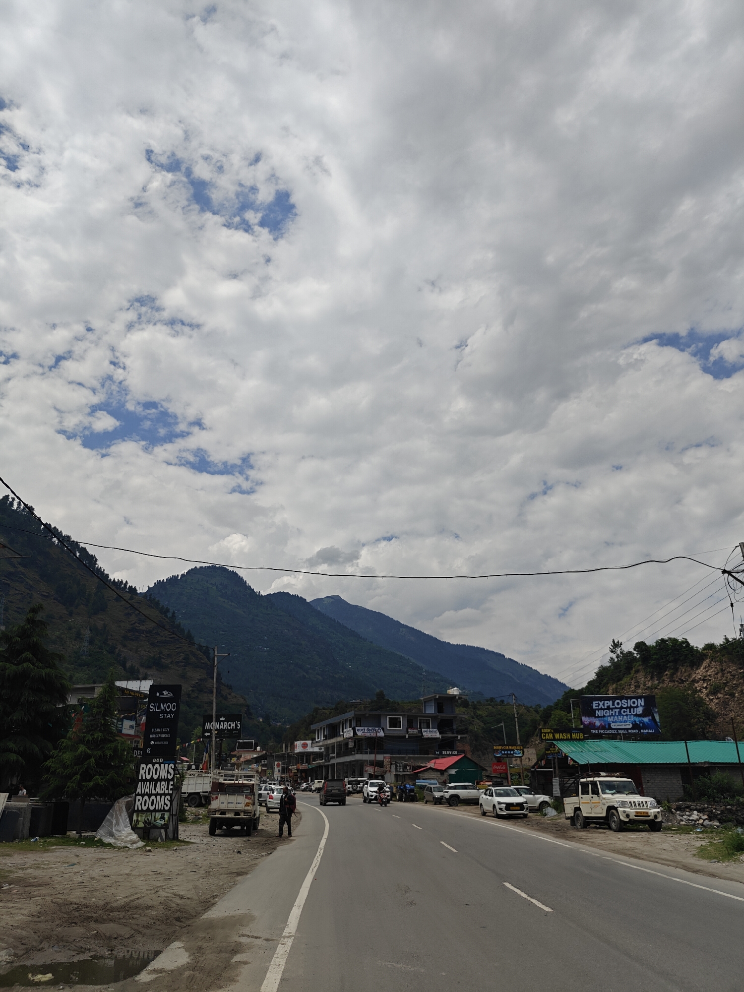 The image presents a daytime view of a road in Patlikuhal, India, winding through a mountainous landscape. The scene is dominated by a wide, paved road with a white shoulder line, extending into the distance where light traffic is visible. On both sides, steep mountains covered in dense green foliage and trees rise dramatically under a predominantly cloudy sky, with some patches of blue peeking through.
Along the left side of the road, a large black and white sign advertises "SILMOO STAY CLEAN & COZY WORDEN ROOMS B&B ROOMS AVAILABLE ROOMS". Further down, a smaller sign indicates "MONARCH'S". On the right, several vehicles are parked, including a white SUV and a white pickup truck, with a person standing near the latter. Commercial buildings line this side of the road, featuring signs such as "BODI'S CAR WASH HUB" and "SERVICE". High on the hillside to the right, a prominent billboard promotes "EXPLOSION NIGHT CLUB MANALI THE PICCADILY, MALL ROAD, MANALI". Utility poles and electrical wires crisscross the upper portion of the frame against the cloudy sky. The overall impression is of a lively mountain town or highway corridor.