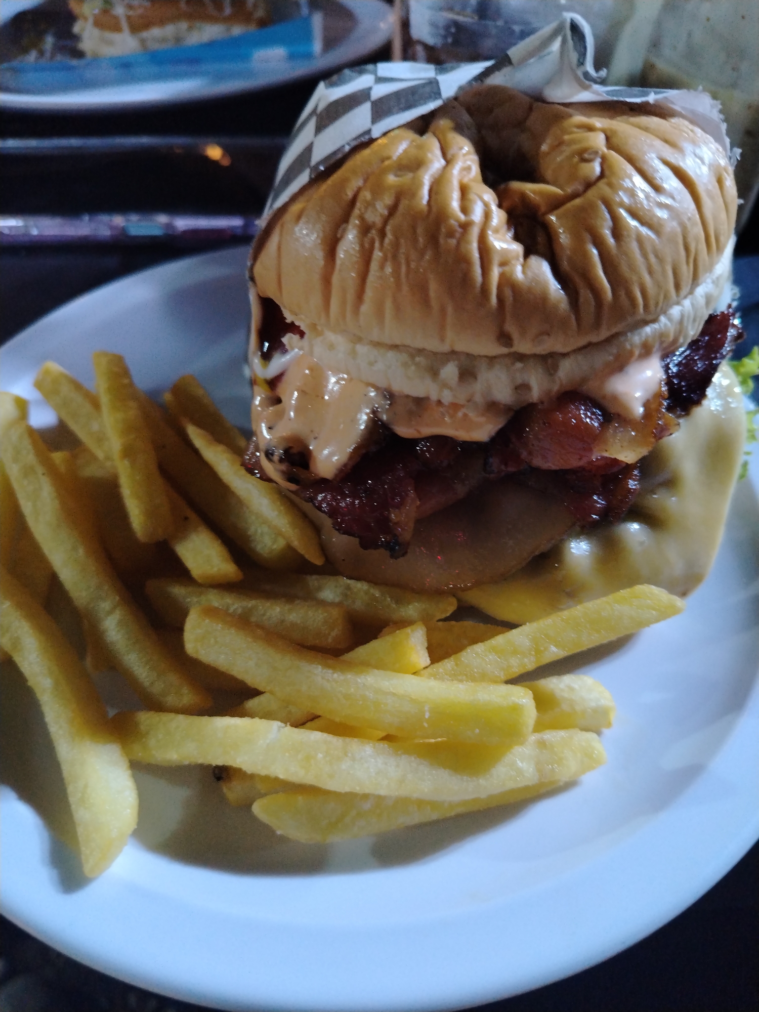The image shows a hamburger and french fries served on a white plate. The hamburger appears to have bacon, cheese, and sauce. A black and white checkered paper wraps the top of the burger. The picture was taken at a restaurant in Tinaquillo, Venezuela. There's another plate with food, possibly another burger, in the background. The setting appears to be indoors, possibly at night.