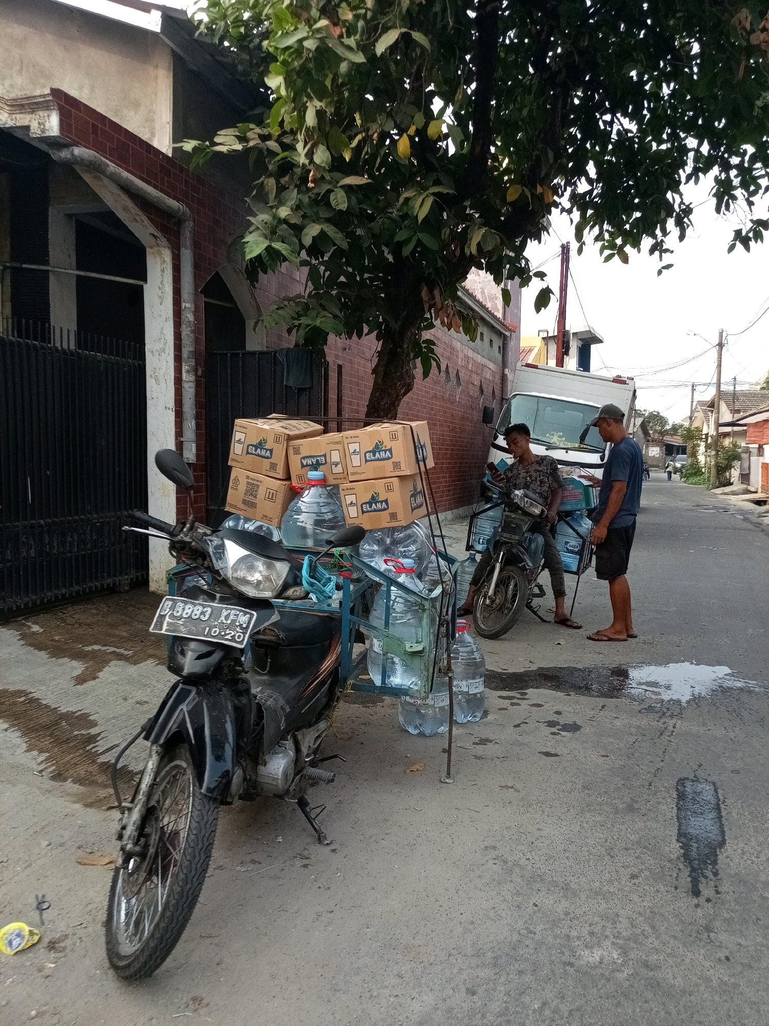 The image depicts a street scene in Bekasi, Indonesia, on a sunny day. A motorcycle, laden with stacks of boxed water and several large water jugs, is parked on the side of a paved road. A man wearing a dark t-shirt and dark shorts, with a cap on his head, stands beside the motorcycle, interacting with another man. The second man, seated on the motorcycle, is wearing a patterned t-shirt and looking at his phone. Behind them, a white truck is parked, and further down the street, buildings and other vehicles are visible. The ground is wet in patches, suggesting recent rain or spillage. The environment appears to be a residential or commercial area with a tree providing shade. The license plate of the motorcycle reads "B 8883 KFM".