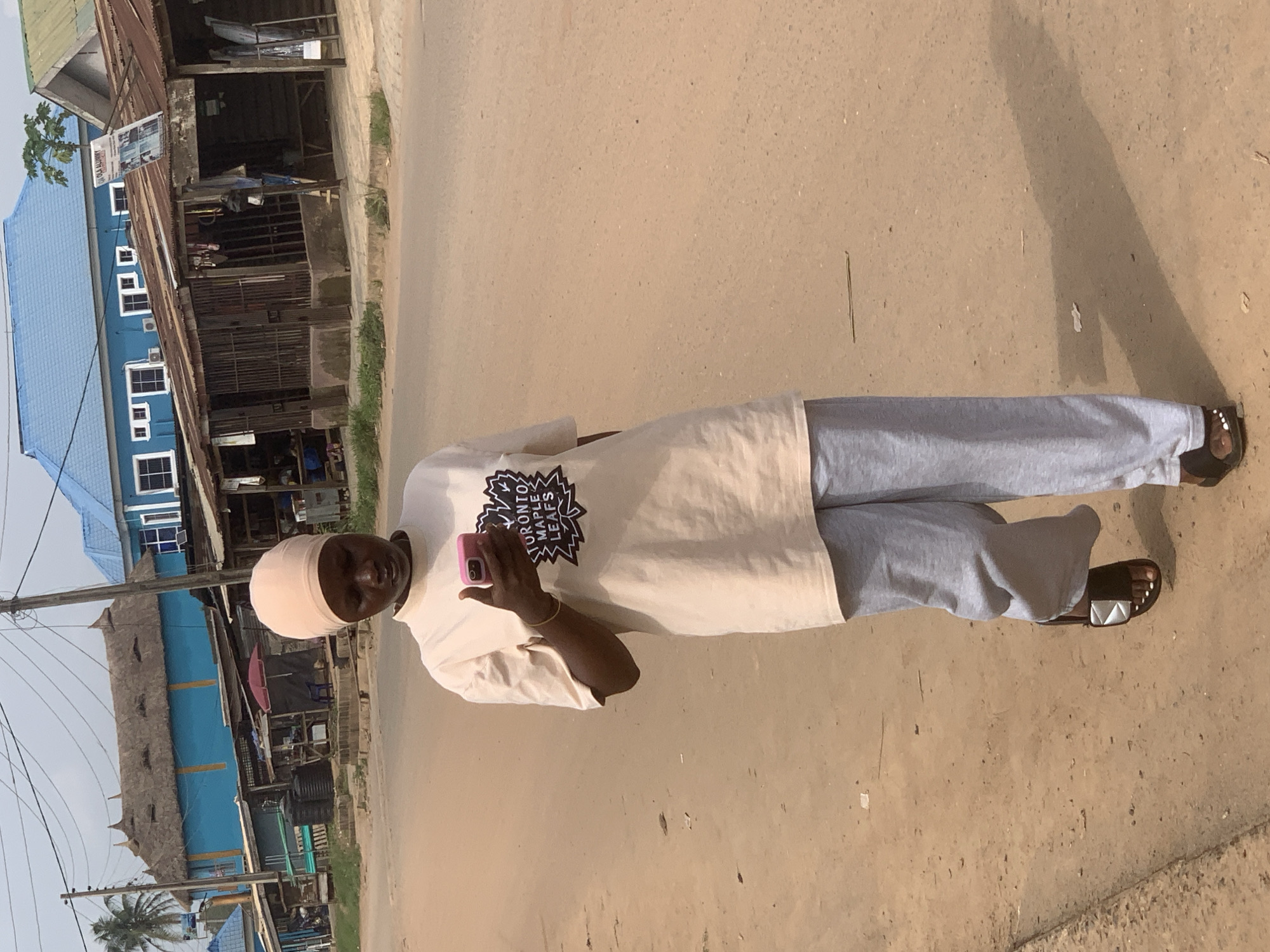 The image shows a person standing in what appears to be a street. The individual is wearing a light-colored head covering, a light tan t-shirt with the Toronto Maple Leafs logo, and grey sweatpants. The person is holding a pink mobile phone.
The background features buildings with corrugated metal roofing, some painted blue. There's also a palm tree visible in the distance. The location appears to be Shagamu, Nigeria, based on the architectural style and environmental characteristics. The street surface is unpaved and dusty.