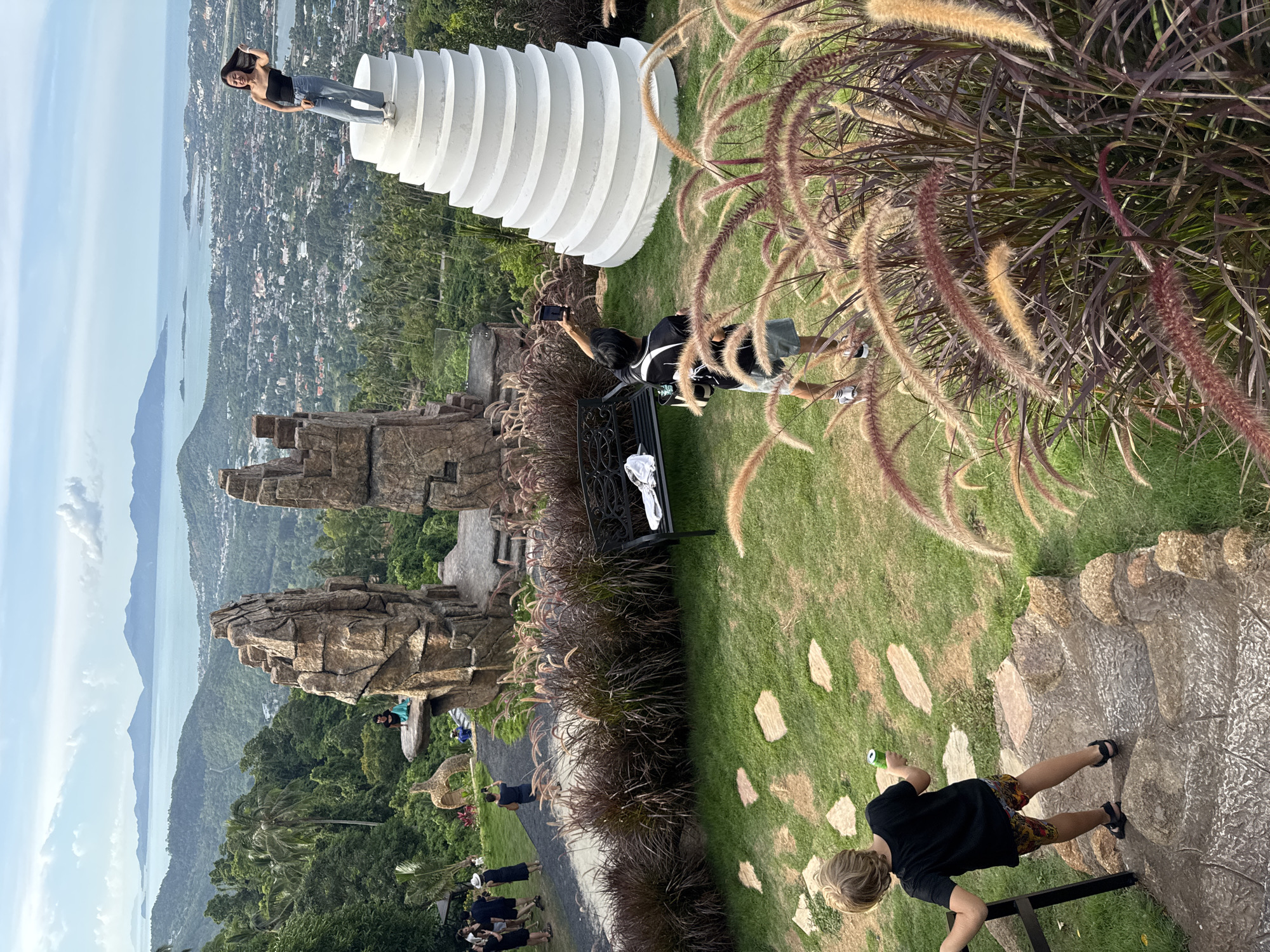 An eye-level, wide outdoor shot captures a scenic view of a city nestled amidst lush green hills, with a body of water and distant islands under a partly cloudy sky. In the foreground, a young boy with blond hair, wearing a black t-shirt and patterned shorts, walks down a stone staircase. To the right, a young person in a black t-shirt and shorts is taking a selfie with a phone, standing on a grassy area with ornamental grasses. Behind them, a woman in a black top and jeans is posing on top of a white spiral structure. Further back, a group of people are walking on a path near large, faux rock formations. A black bench with a white cloth is visible on the grass to the left of the spiral structure. The environment appears to be a viewpoint or park offering panoramic vistas.