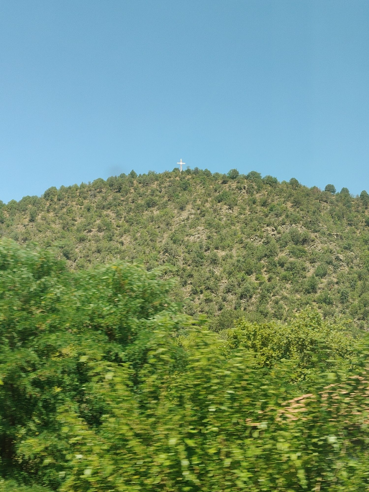 A white cross stands atop a sparsely wooded, rocky hill under a clear blue sky. The foreground is filled with blurred green foliage, suggesting movement. The scene is likely located near the town of Kresna, Bulgaria, given the context of the image.