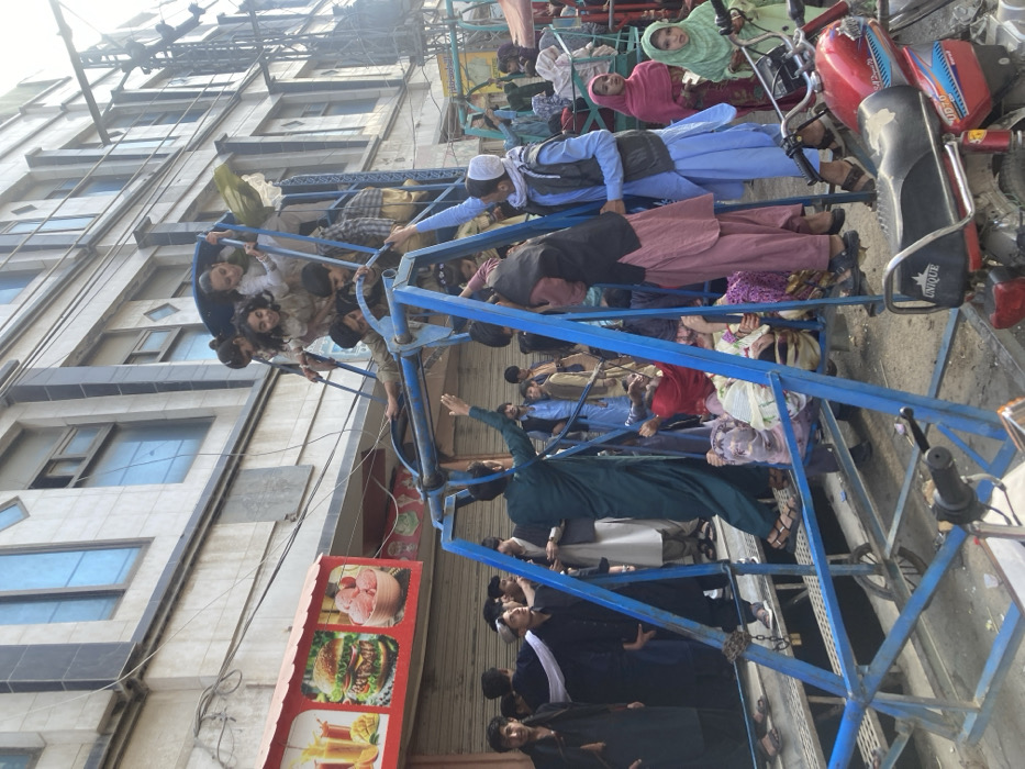 This image depicts a bustling outdoor scene in Quetta City Tehsil, Pakistan, under clear daylight conditions. The central focus is a crowded, blue metal swing set, appearing to be a portable amusement structure. Approximately 15 to 20 individuals, predominantly children ranging in age from young to adolescent, are actively engaged with the swing set. Multiple children are seen standing on the swing seats, some smiling, while others are seated or climbing the blue metal frame.
Several adult males are present near the swing set. One male, wearing a dark green shalwar kameez, stands with his arms raised, seemingly interacting with the children on the upper part of the structure. Another male, dressed in a light blue shalwar kameez and a dark vest, stands observing the activity. A larger group of individuals, primarily males in traditional Pakistani attire, forms a dense background crowd, indicating a public gathering or market street. Several women and younger girls, some wearing head coverings, are also visible among the bystanders.
In the foreground, the rear section of a red motorcycle, featuring a "UNIQUE" branded seat and "CD 70" visible on its side panel, is partially parked next to the swing set. The background consists of a multi-story urban building with a light-colored facade and multiple rectangular windows, some tinted blue. To the left, a red and white food stall sign is visible, displaying stylized images of juice, a hamburger, and ice cream. Overhead electrical wires are evident across the scene. The ground surface appears to be unpaved or a worn asphalt.