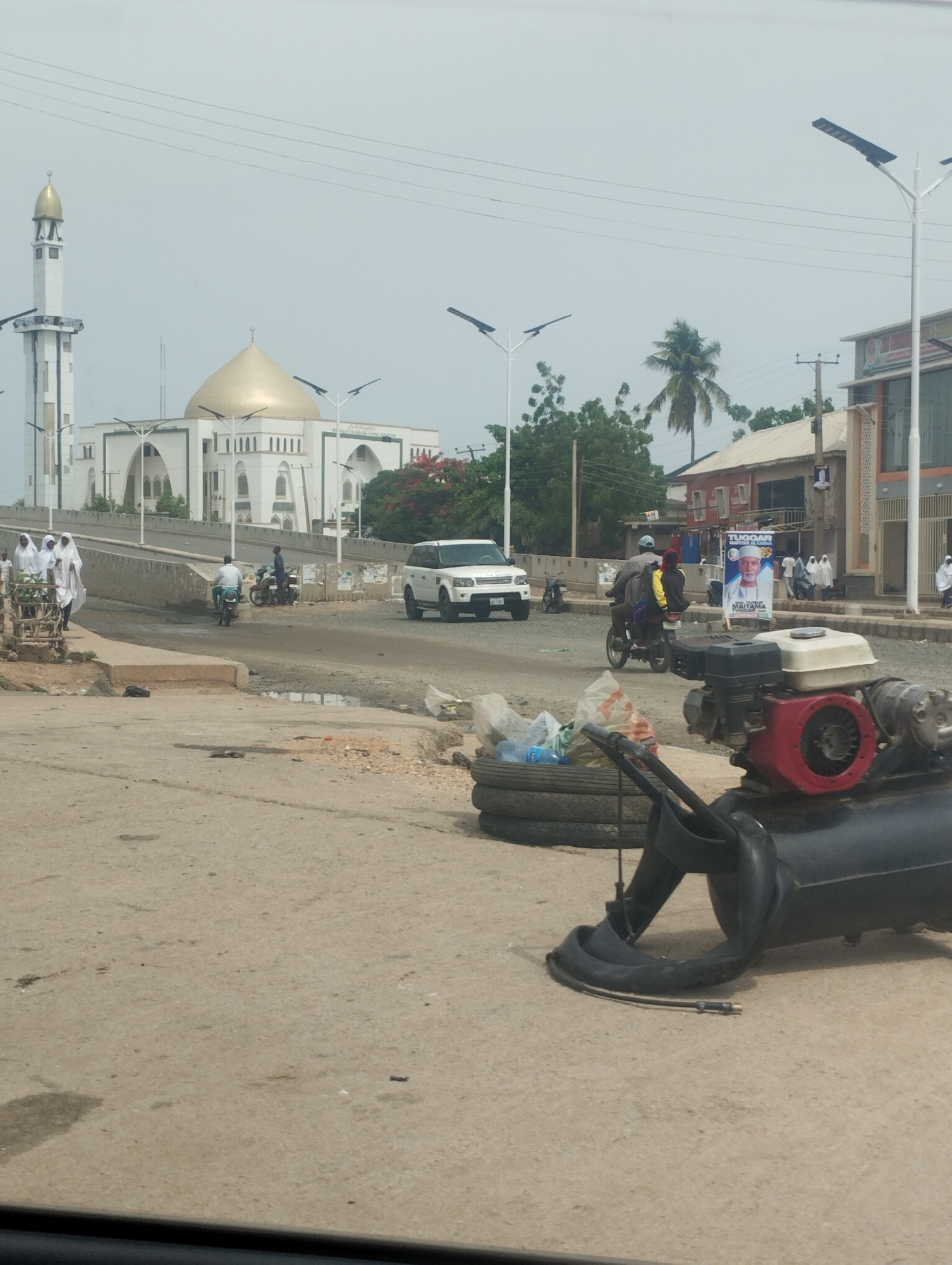 The image depicts an urban street scene in Bauchi, Nigeria. In the background stands a large, white mosque with a golden dome and minaret. In the middle ground, there is a raised road with a white SUV driving on it. A group of people dressed in white robes are seen walking near the bridge. Motorcycles are also visible on the road. In the foreground, a black air compressor sits on the pavement next to a stack of tires. A political poster is affixed to a building in the background. Overhead power lines stretch across the sky.