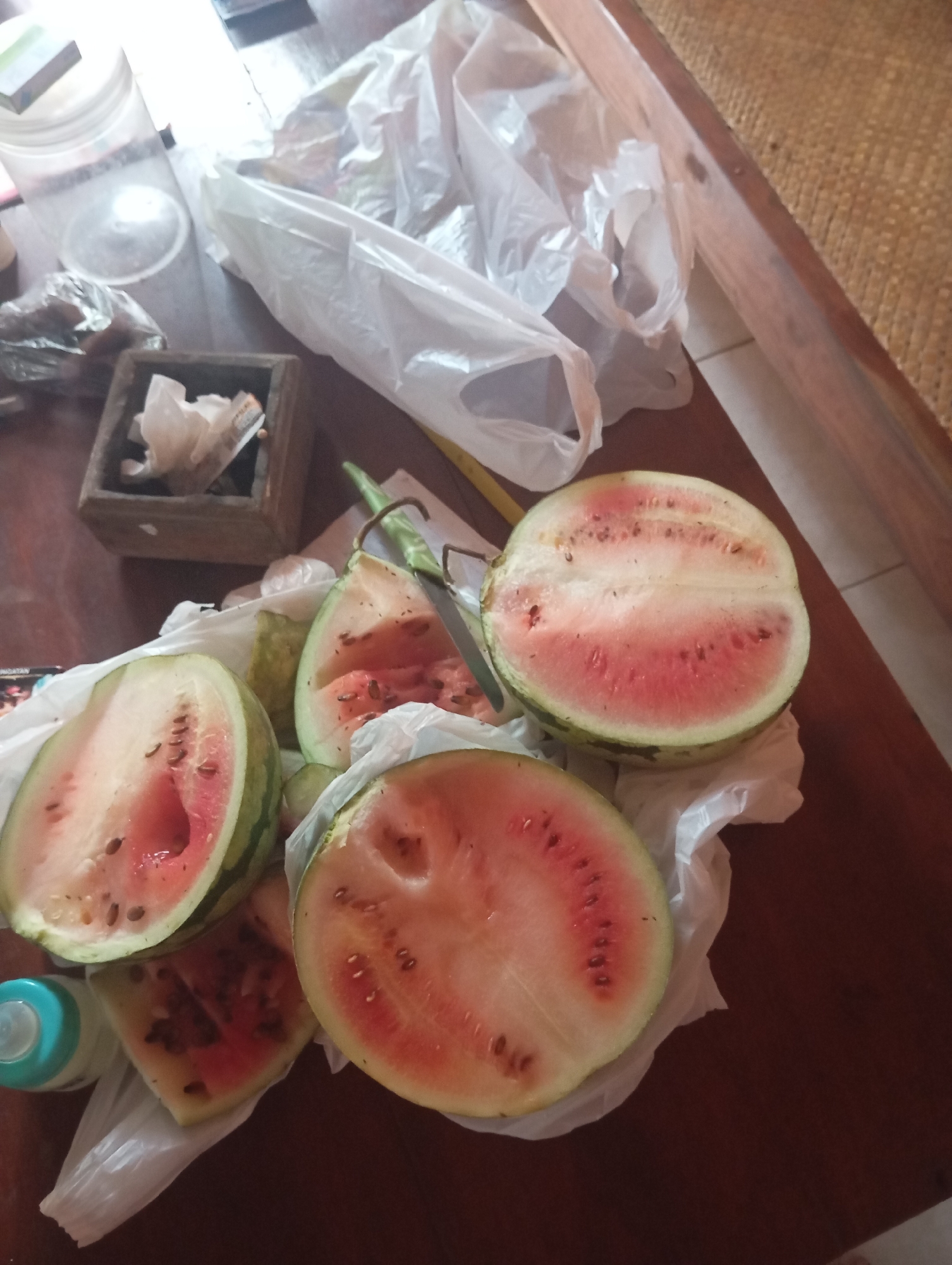 On a wooden table in Kalialang, Indonesia, several cut watermelons are displayed. The watermelons are mostly pink with visible seeds and are arranged on white plastic bags. There are also other items on the table, including a wooden box with some white objects inside, a plastic container, and a blue baby bottle. The overall scene suggests a casual setting, possibly a home or outdoor area, with the watermelons prepared for consumption.