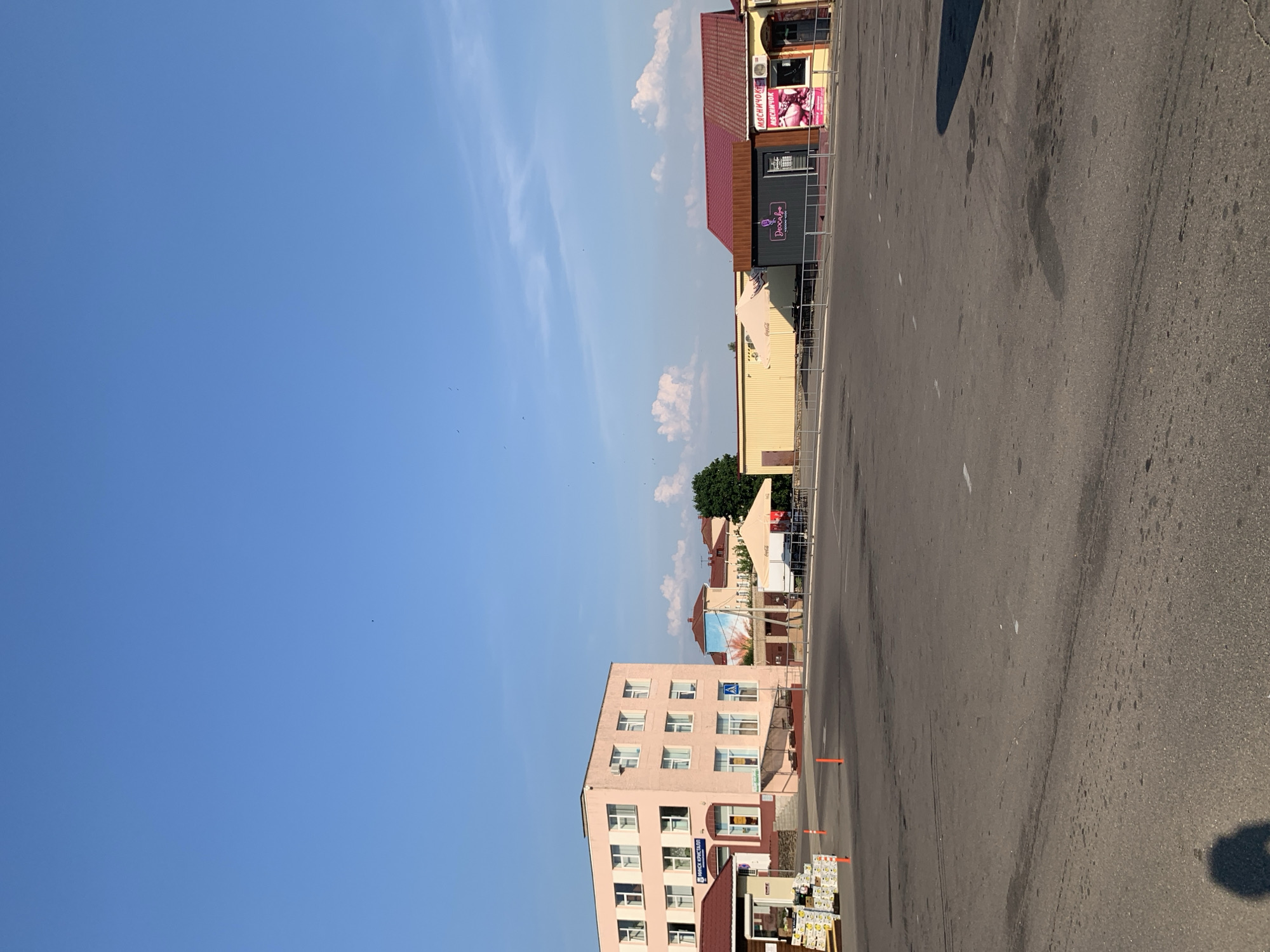 The image depicts an outdoor scene in the city of Kapyĺ, Belarus, likely a public square or market area. In the background, several buildings are visible. A prominent pale pink, multi-story building with numerous windows stands on the left. To its right, a yellow, single-story building with a red-tiled roof appears to house small businesses, one with a red awning and a sign that reads "МЯСНИЧОК". A dark grey structure with a pink logo "Deoxa.by" is situated between the yellow building and the pink one, also under a red roof. Several outdoor umbrellas and tables are set up in front of these structures, suggesting an area for seating or outdoor commerce. The foreground is dominated by a paved asphalt surface, possibly a parking lot or pedestrian walkway, marked with faint white lines. A shadow of a person is visible in the lower left corner. The sky above is a clear, bright blue with a few scattered white clouds and some very small dark specks, possibly birds. Orange traffic cones are placed on the pavement near the pink building. The overall impression is that of a typical urban setting on a sunny day.