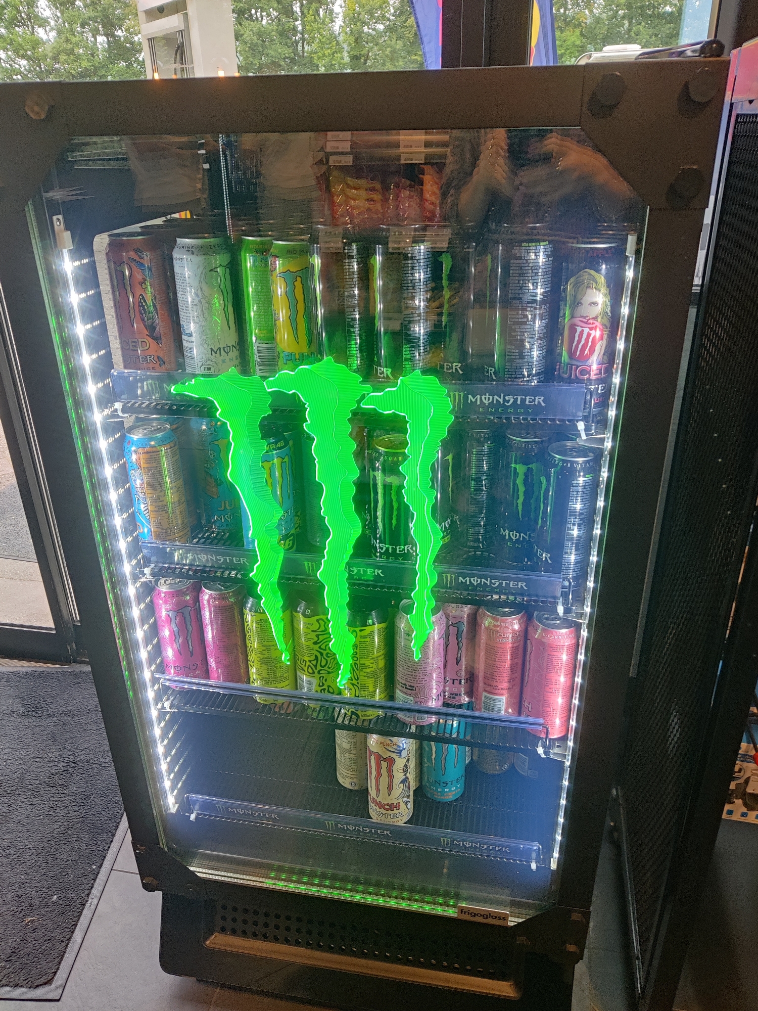 In Eijsden, Netherlands, a tall, illuminated beverage cooler displays rows of Monster Energy drinks. The cooler's glass door reveals multiple shelves stocked with cans of various Monster flavors, including green, pink, and blue varieties. A prominent, bright green, stylized Monster logo is affixed to the front of the cooler, illuminated by internal LED lights. The scene is captured from a slightly angled, frontal perspective, with the cooler positioned indoors near an entrance. Outside, through the glass, glimpses of trees and a building facade are visible, suggesting an outdoor environment adjacent to the indoor setting of the cooler. The floor in front of the cooler is covered by a dark grey mat.