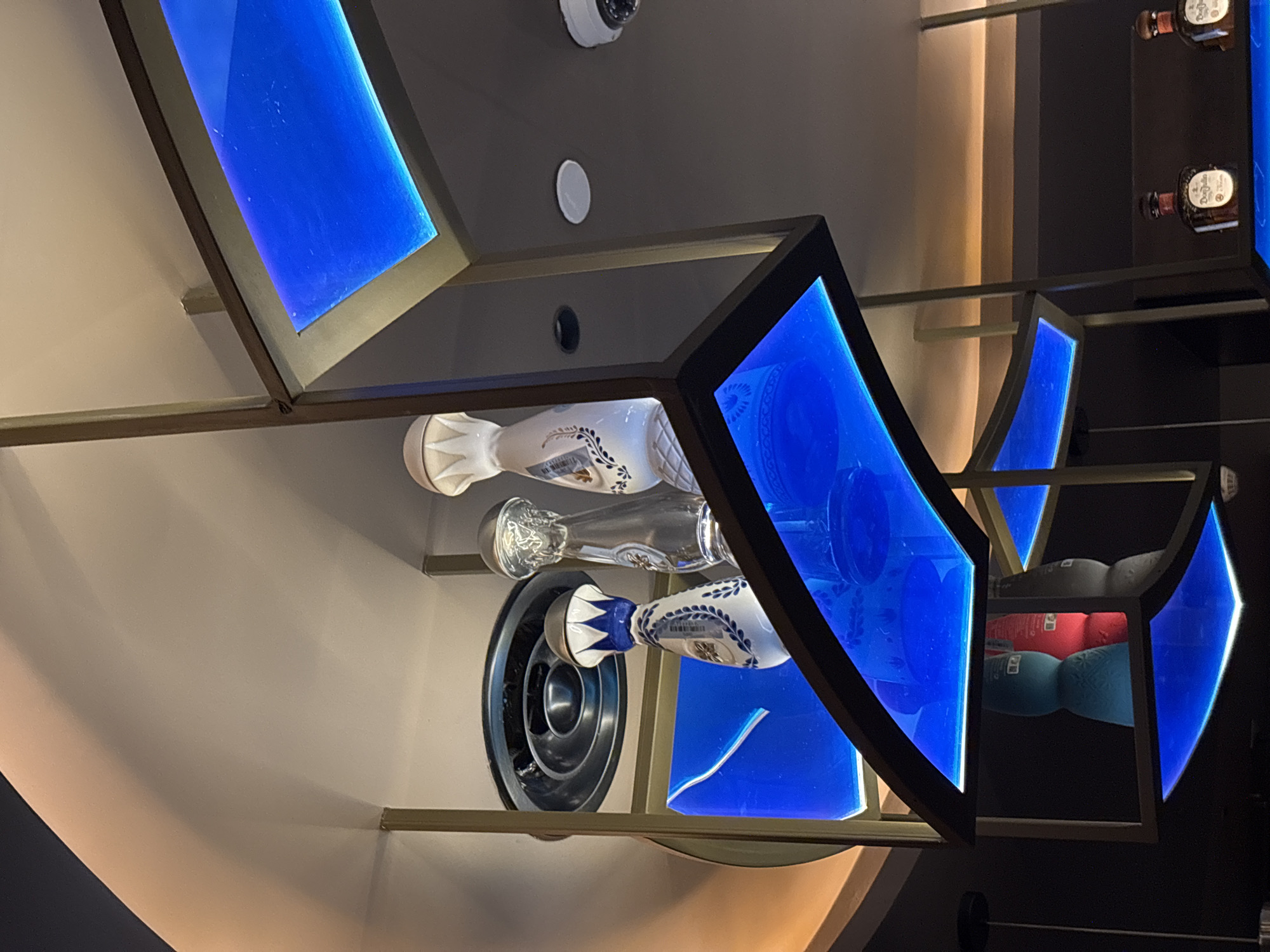 The image shows a display of decorative bottles on multiple shelves in what appears to be a bar or restaurant. The shelves are illuminated with blue LED lights, creating a vibrant ambiance. On the top shelf, there are three bottles: one is white with blue decorative patterns, another is clear glass, and the third is white with a ribbed texture. Below this shelf, on a blue illuminated surface, are two more bottles, one blue and one red, partially obscured by the shelf above. Further down, on a lower shelf also illuminated by blue light, are two brown bottles with red labels. The overall setting is modern and dimly lit, with warm lighting visible on the ceiling in the upper portion of the image. There are no people visible in the frame. The location is identified as Amman, Jordan.
