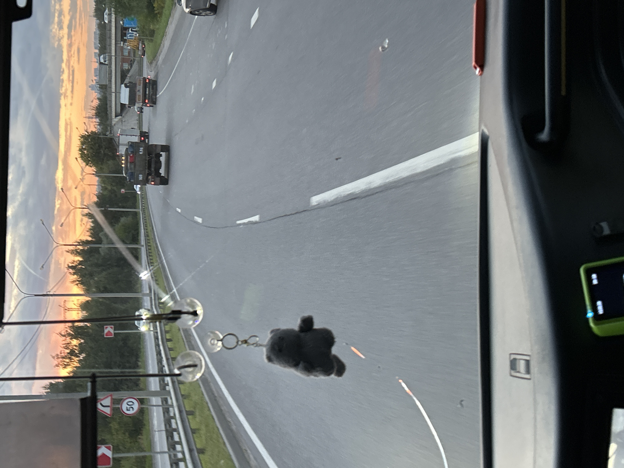The image shows a view from inside a vehicle, looking out at a highway during sunset. In the foreground, a small, dark gray stuffed bear keychain hangs from the rearview mirror. The road ahead is paved, with white lane markings. Several vehicles are visible on the highway, including trucks and cars. The sky is filled with warm, orange and yellow hues, indicating the sun is setting. Trees line the side of the highway. Traffic signs are visible on poles, including a speed limit sign indicating 50. The dashboard of the vehicle is visible at the bottom of the image.