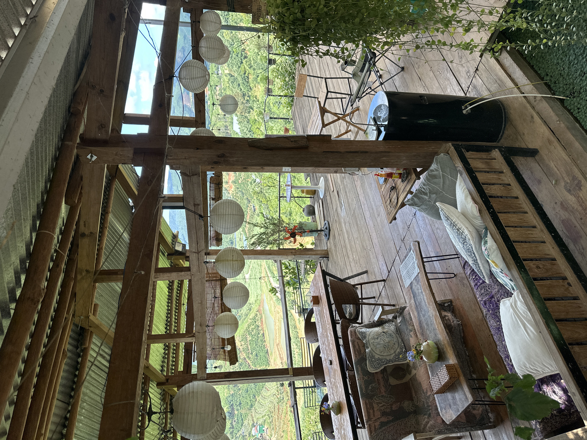 This is a top-down, rotated shot of a rustic, open-air cafe or seating area in Thôn 3, Vietnam. The setting features a wooden deck overlooking a lush, green landscape with terraced fields and water features, indicative of a rural or agricultural area. Hanging paper lanterns in white and spherical shapes are a prominent decorative element, strung across the wooden beams that support a corrugated metal roof on one side and open to the sky and view on the other.
The main subject is the seating area, which includes a long wooden table with several wicker chairs around it. Decorative pillows with intricate patterns are placed on the table and on a bench or seating area to the right. A small vase with flowers and a woven box sit on the table. A black cylindrical object, possibly a water cooler or a decorative drum, is positioned on the deck further back.
The time of day appears to be daytime, with bright sky visible through the open sections of the roof. The weather seems pleasant, suitable for an outdoor setting. The overall ambiance is relaxed and natural, with the greenery and open design contributing to a serene atmosphere. There is no visible text in the image.