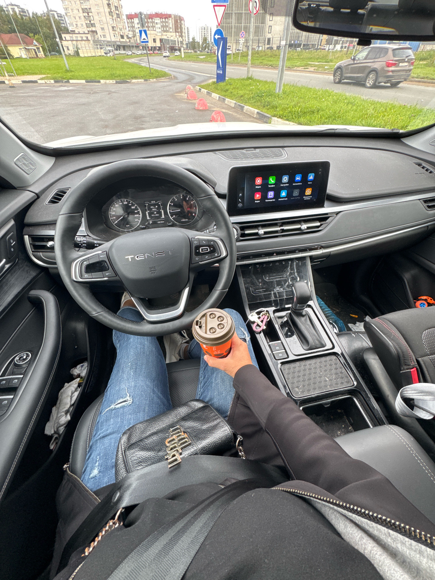 The image captures the interior of a car, likely a Chery Tiggo 7 Pro, viewed from the perspective of the driver. The driver, wearing distressed blue jeans and a black jacket, is holding a disposable coffee cup. A black leather handbag with gold lettering rests on their lap. The car's dashboard features a large touchscreen infotainment system displaying various icons, a digital instrument cluster, and a multi-function steering wheel. The gear shifter, center console, and part of the passenger seat are also visible. Outside the car, through the windshield, a street scene unfolds with apartment buildings, other vehicles, and traffic signs, suggesting an urban setting. The location is identified as Novoselye, Russia.
