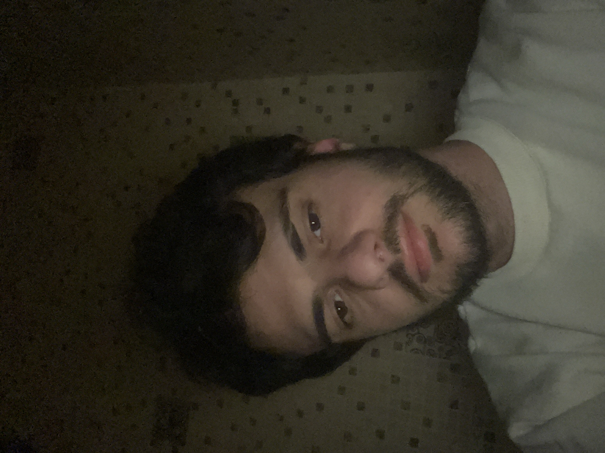 The media file presents a dark, low-light image of a man lying down, with the photograph rotated 90 degrees clockwise. The man, who appears to be an adult with dark hair and a beard, is positioned on the left side of the frame, looking towards the top right. He is wearing a plain white t-shirt or top. His eyes are open, reflecting a faint light source, and his expression is somewhat neutral or pensive. The background consists of a dark, textured surface, possibly a carpet or patterned blanket, featuring a subtle repeating pattern of small, square-like shapes. The overall lighting is very dim, suggesting an indoor setting at night or in a poorly lit room. No specific location cues for Tbilisi, Georgia, are visible within the image itself, though this is the provided context. There is no visible text. The man appears to be resting or relaxing.
