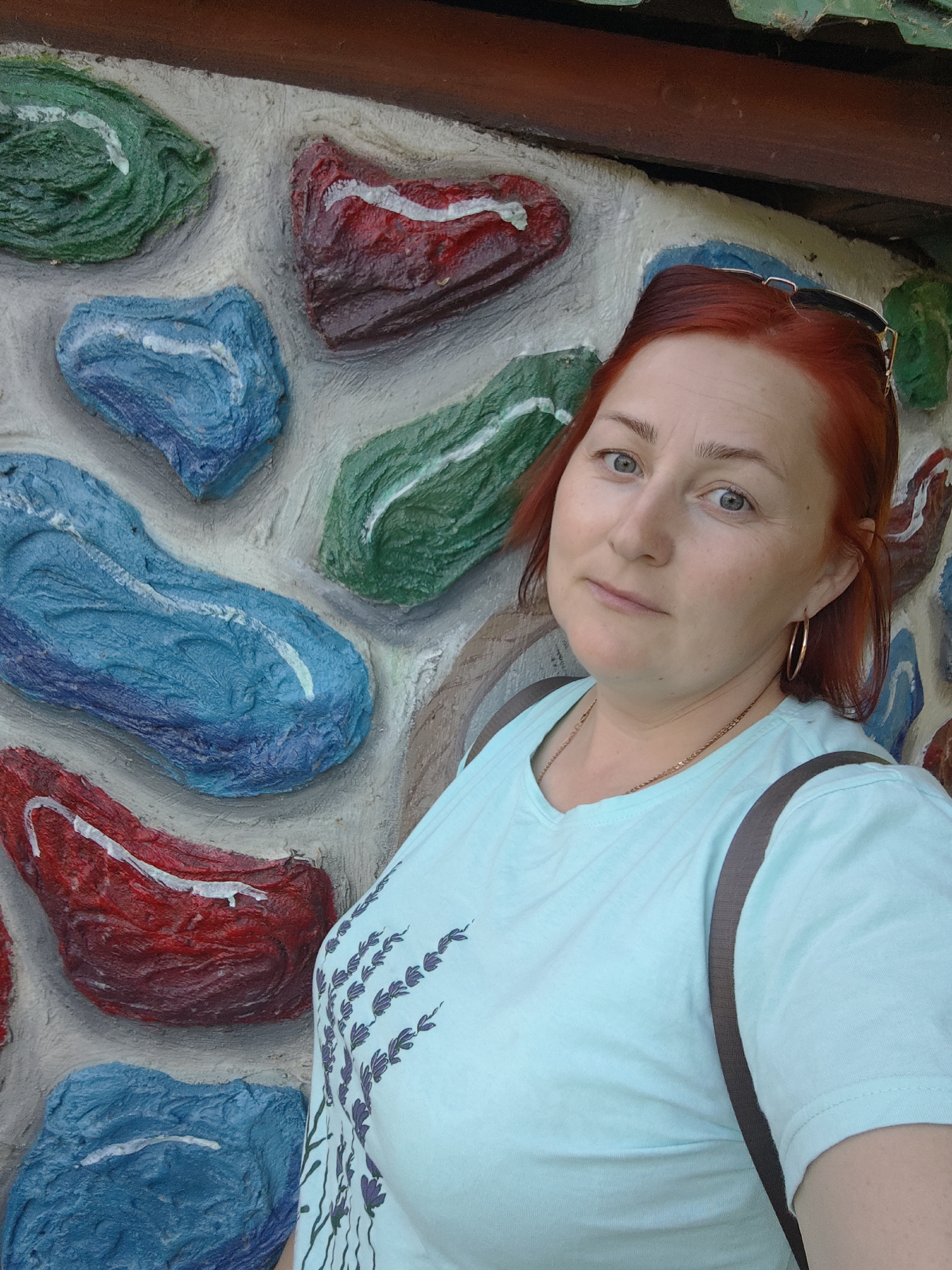 A woman with red hair is standing against a textured wall comprised of faux rocks painted in various bright colors including blue, red, and green. The wall also has a brown strip at the top. The woman is wearing a light blue t-shirt with a floral design on the front. She has gold hoop earrings and a gold chain necklace. Sunglasses are perched on top of her head. She appears to be wearing a bag with a gray strap over her shoulder. The location is Chaikovsky, Russia.