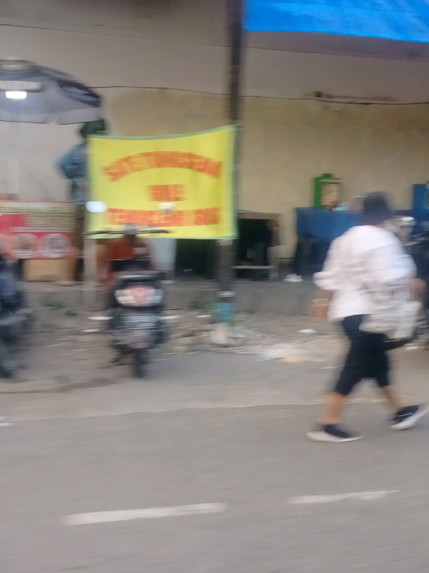 The image depicts a street scene in East Jakarta, Indonesia, characterized by motion blur. In the foreground, a person wearing a white patterned shirt and dark pants walks from left to right. They are carrying a bag. In the midground, a motorcycle is parked. Behind the motorcycle, a yellow banner with red text is visible. The background shows a building with a blue awning, a white tent, and various other structures, suggesting a commercial or street market area. The ground is paved, and there are some scattered debris. The overall impression is one of a bustling urban environment captured in motion.