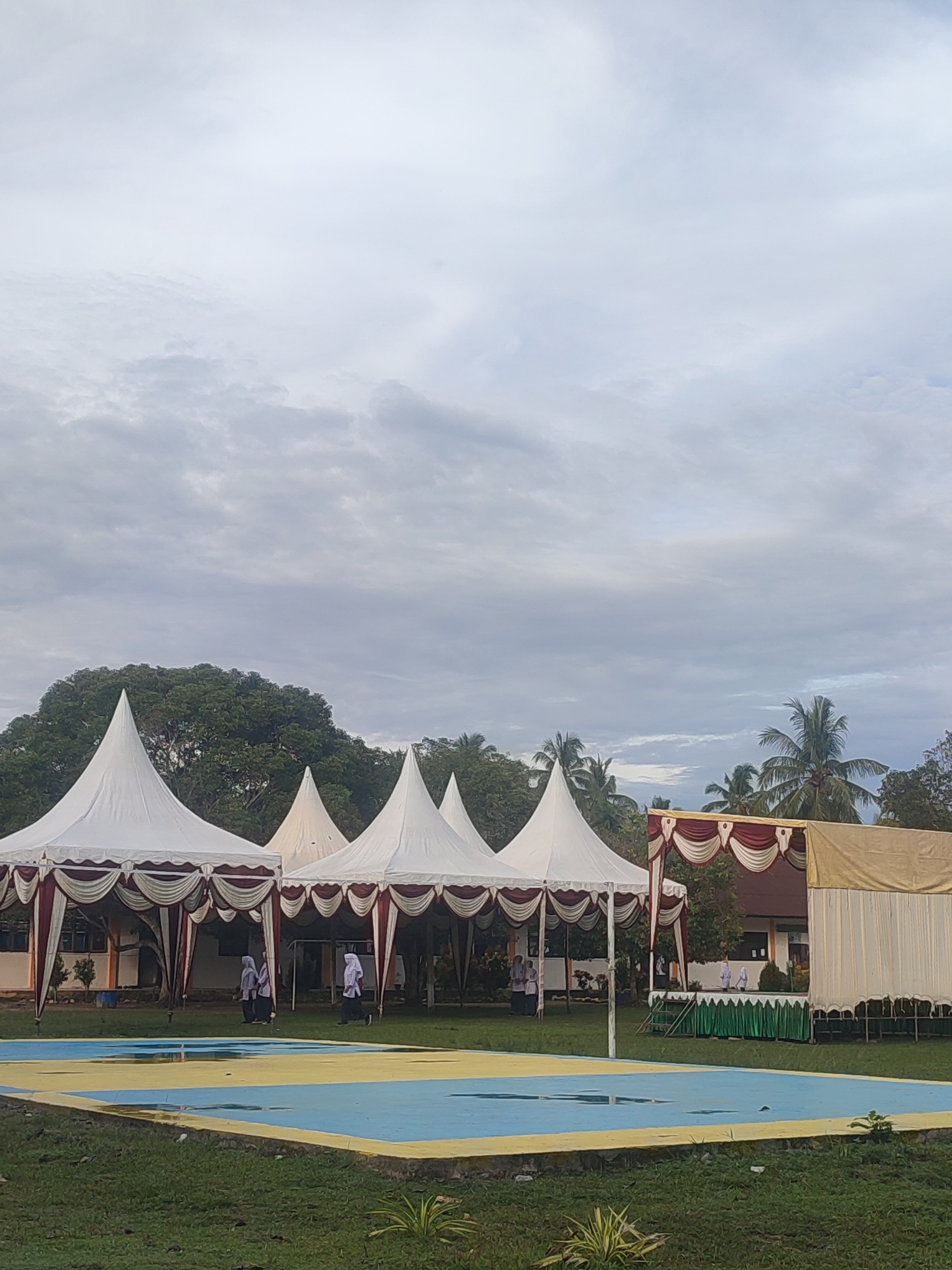 The image shows a scene outdoors under a cloudy sky. Several white tents with red and cream-colored draping line the edge of what appears to be a schoolyard or public event space in Blangriek, Indonesia. In the background are green trees, including palm trees, and low-rise buildings. In the foreground, there is a blue and yellow painted surface, possibly a sports court, surrounded by green grass. A small stage draped with fabric is positioned towards the right. Several people in white outfits are walking around between the tents and the buildings.