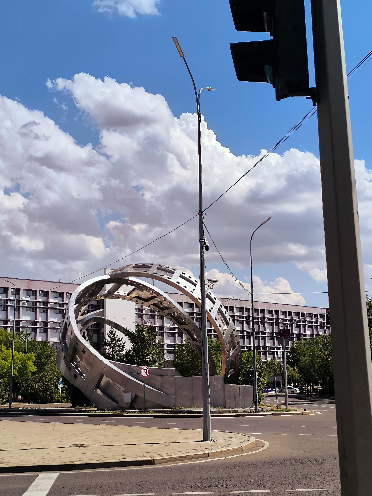The image shows a large, metallic, spherical sculpture located in the city of Karaganda, Kazakhstan. The sculpture, composed of intersecting metal rings and panels, is situated in a roundabout at an intersection. Behind the sculpture, a long, multi-story building with a grid-like facade and numerous windows is visible. Trees and greenery surround the sculpture and the base of the building. A traffic light pole with a traffic signal and a street lamp are present in the foreground and midground. The sky is blue with scattered white clouds. The ground is paved with asphalt and concrete, indicating a public urban space. There are no people visible in the image.