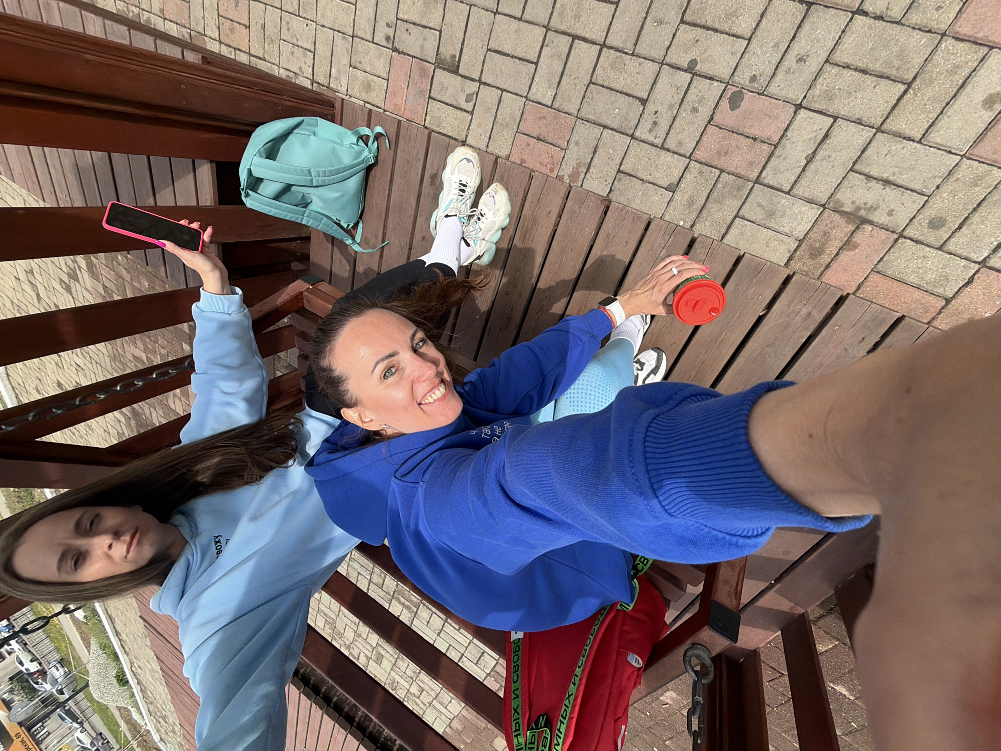 Two women pose for a selfie on a wooden structure in Birobidzhan, Russia. One woman, wearing a blue hoodie and red pants with green accents, holds a red-lidded cup and smiles at the camera. The other woman, wearing a light blue hoodie, holds up a pink-cased cell phone. A light blue backpack rests nearby. The background shows a paved area and a cityscape.