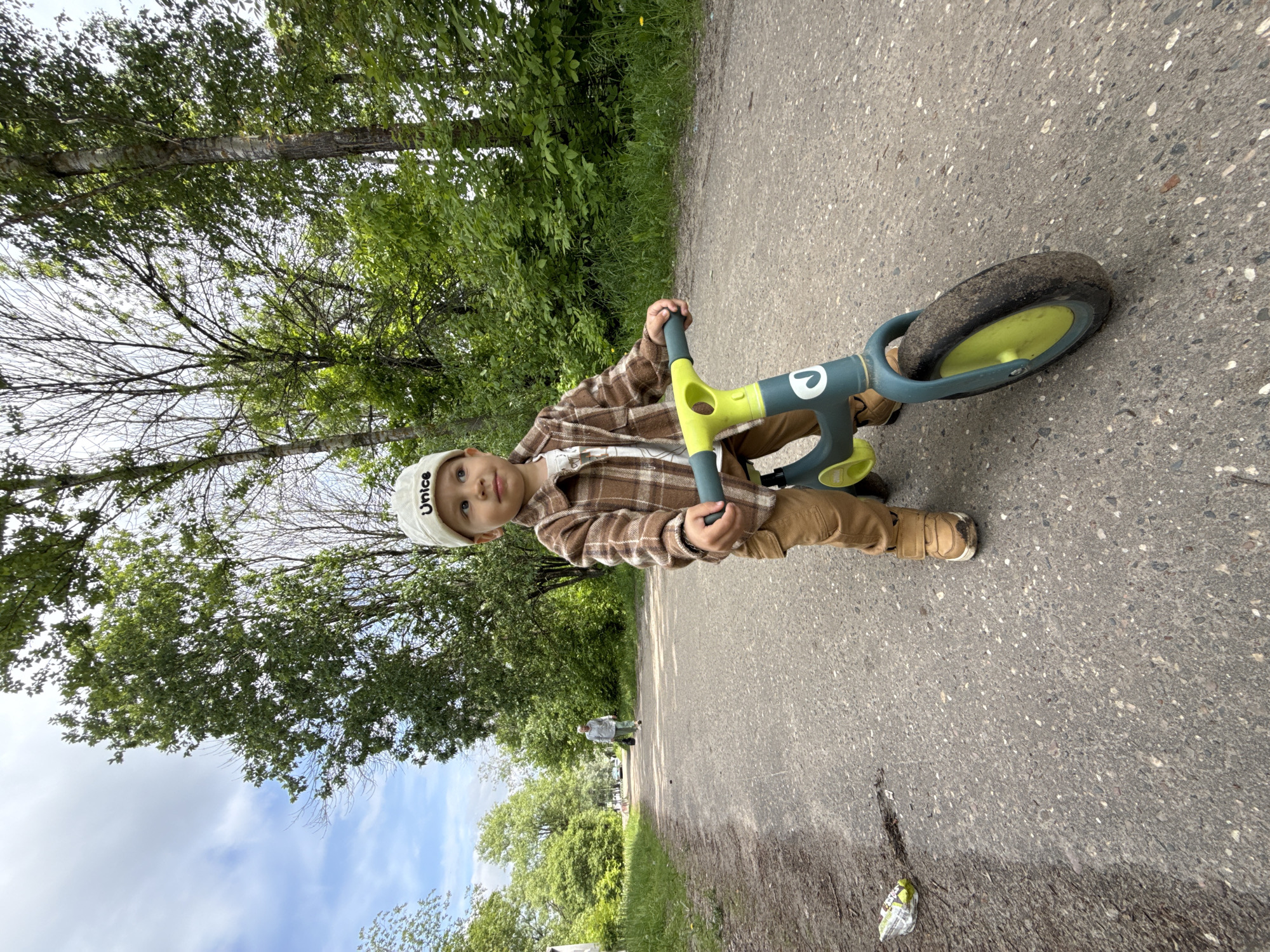 A toddler wearing a brown plaid shirt, khaki pants, and tan shoes is riding a balance bike on a paved path. The child is also wearing a white cap. The balance bike has green and gray components and a single tire. The scene is outdoors, with trees and foliage visible on the left side of the path. A person is walking in the distance on the same path. Trash is on the ground in the foreground. The location is Заволжье, Russia.