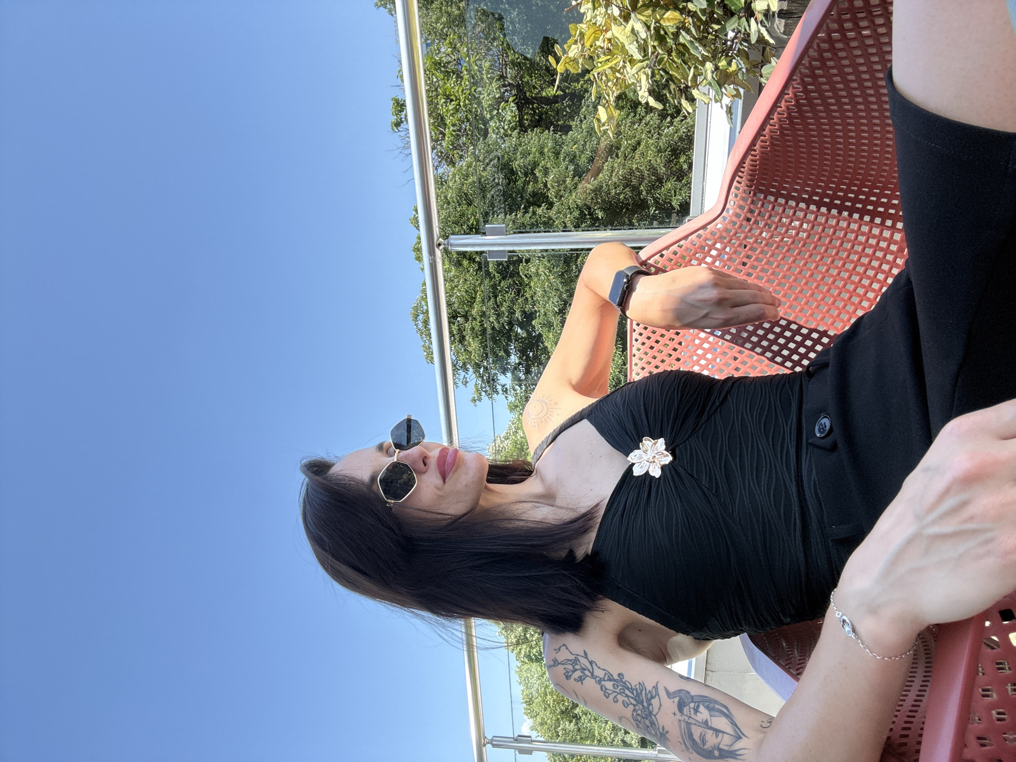 A woman is seated in a red, perforated metal chair outdoors. She wears black clothing and sunglasses. A silver flower brooch is pinned to her top. Her left arm has a tattoo, and she wears a silver bracelet on her right wrist. She has dark hair. In the background, there's a clear blue sky, trees, and a glass railing. The location is in Pleven, Bulgaria.