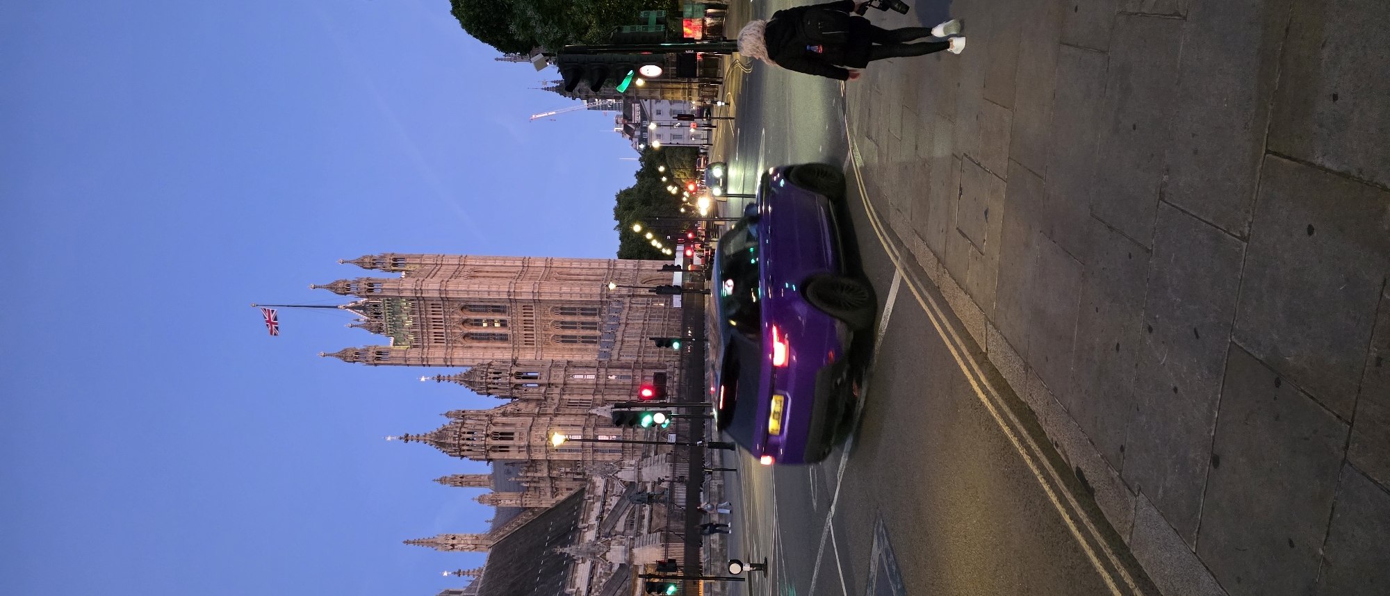 The image captures a view of the Palace of Westminster in London, United Kingdom, during twilight. A purple Range Rover is in motion, turning at an intersection. A woman with blonde hair, wearing a black jacket and leggings, walks on the sidewalk carrying a camera. The Palace of Westminster, a Gothic Revival building, dominates the background, with the Union Jack flag flying from its tower. Traffic lights are visible, with some illuminated red. The street is paved with stone, and there are streetlights lining the road. The sky is a deep blue, indicating the transition from day to night.