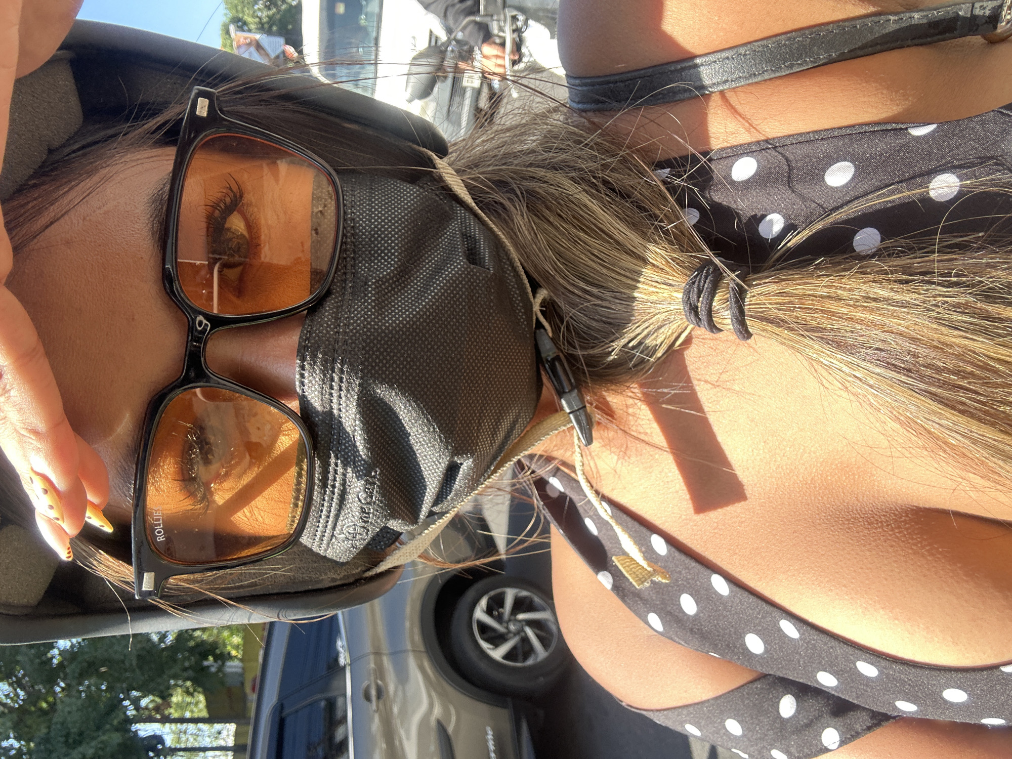 A woman, wearing a black helmet and sunglasses with orange lenses, is riding on a vehicle in Denpasar, Indonesia. She has a black face mask on, covering her nose and mouth. Her hair is tied in a ponytail with a black hair tie. She is wearing a black and white polka dot top. The background shows a street scene with a car and a motorcycle. The lighting suggests it is daytime with bright sunlight.